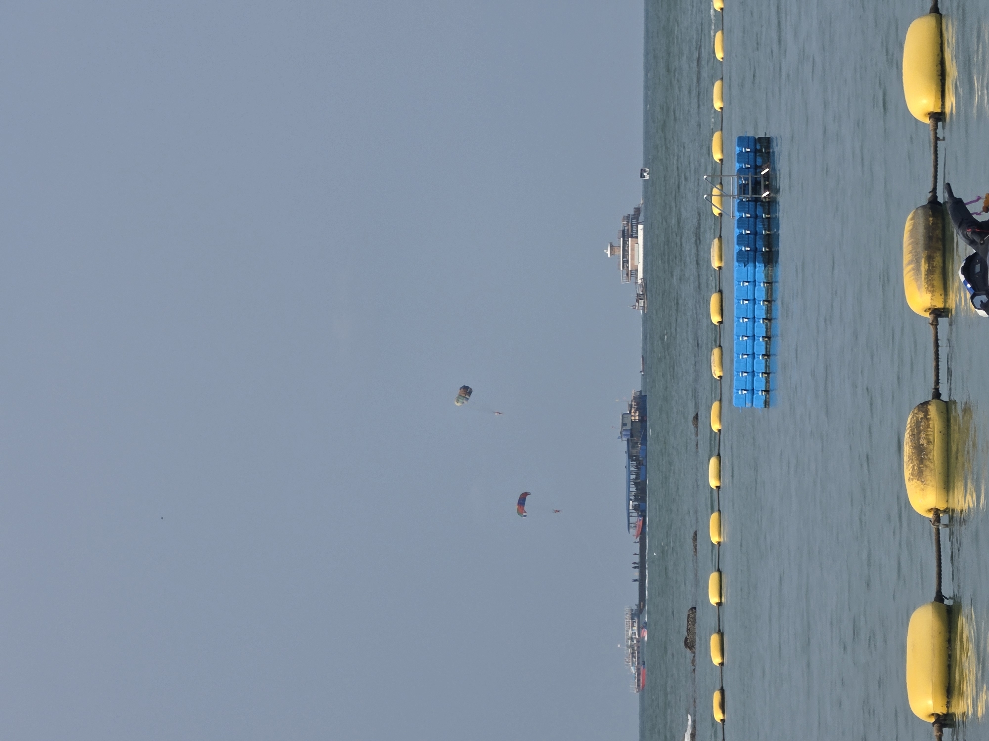 The image displays a vibrant daytime scene of coastal waters in Pattaya City, Thailand, characterized by a clear, light blue sky and a calm, blue-green sea.
In the foreground on the right, a prominent chain of large, bright yellow buoys floats vertically on the water, connected by a dark rope or chain. These buoys demarcate an area within the water. Partially visible behind the uppermost buoy, a person wearing dark clothing appears to be holding onto the buoy line. To the left of this buoy chain, a rectangular blue floating platform or modular dock is present on the water's surface.
Further out in the mid-ground and background, several boats and ships are visible. These include a larger white vessel, possibly a ferry or cruise ship, and other smaller boats, suggesting marine activity or leisure cruises in the distance.
High in the clear sky, two colorful parasailers are engaged in watersports. One parasailer, higher and further away, features an orange, blue, and white canopy. The other, closer and slightly lower, has a distinctive red, blue, and black parachute. These activities suggest a popular tourist destination.
The scene captures recreational activities, with the buoys potentially outlining a swimming or activity zone, and the parasailers adding a dynamic element to the tranquil waterscape. The time of day is clearly daytime, with bright, even lighting. The weather appears fair with clear skies and calm water. There is no visible text within the image. The overall impression is one of leisure and activity typical of a popular seaside resort.
