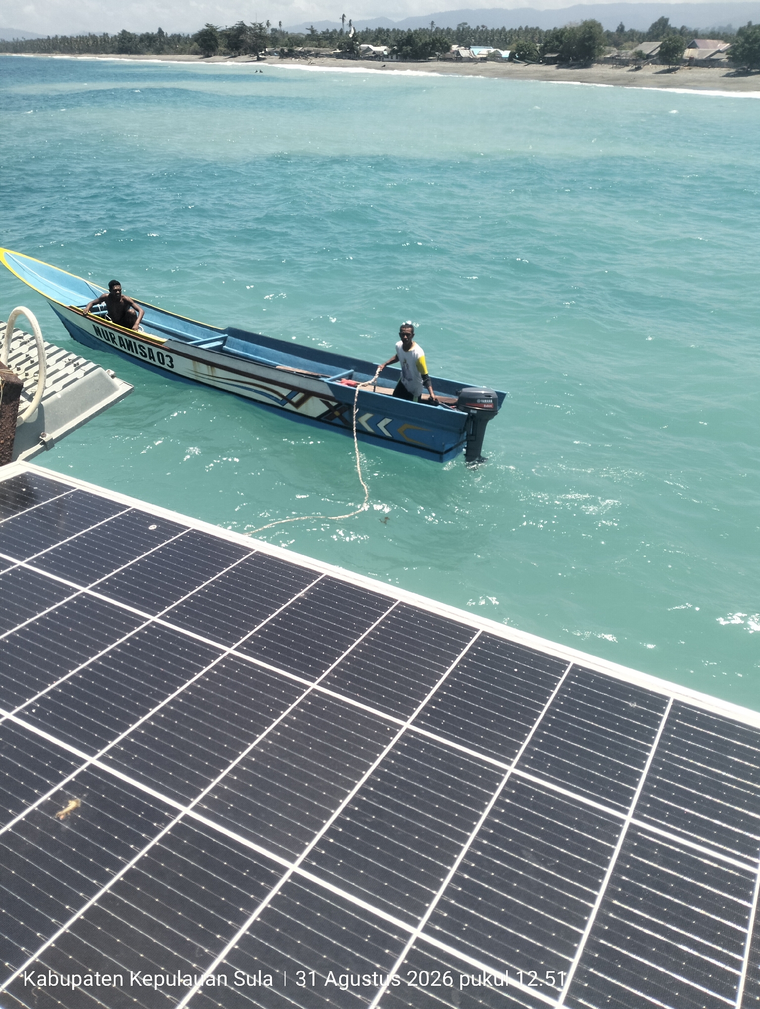 In Pihingtado, Indonesia, a serene ocean scene unfolds with a boat docked near a solar panel array. The boat, named "NURAMISA03", carries two individuals. One man, shirtless and dark-skinned, sits towards the front of the boat. Another man, wearing a white shirt with yellow accents and sunglasses, stands at the stern, operating a Yamaha outboard motor. The boat is tied to a pontoon, and a rope extends from the boat to an unseen point. The water is a clear, turquoise hue, with gentle waves reflecting the bright sunlight. In the background, a sandy beach meets a lush green treeline, dotted with houses. The sky is a pale blue, with a few wispy clouds. The foreground is dominated by a section of solar panels, their dark surfaces glinting in the sun, suggesting a modern infrastructure element in this coastal setting. The timestamp "Kabupaten Kepulauan Sula | 31 Agustus 2026 pukul 12.51" is overlaid on the bottom of the image.