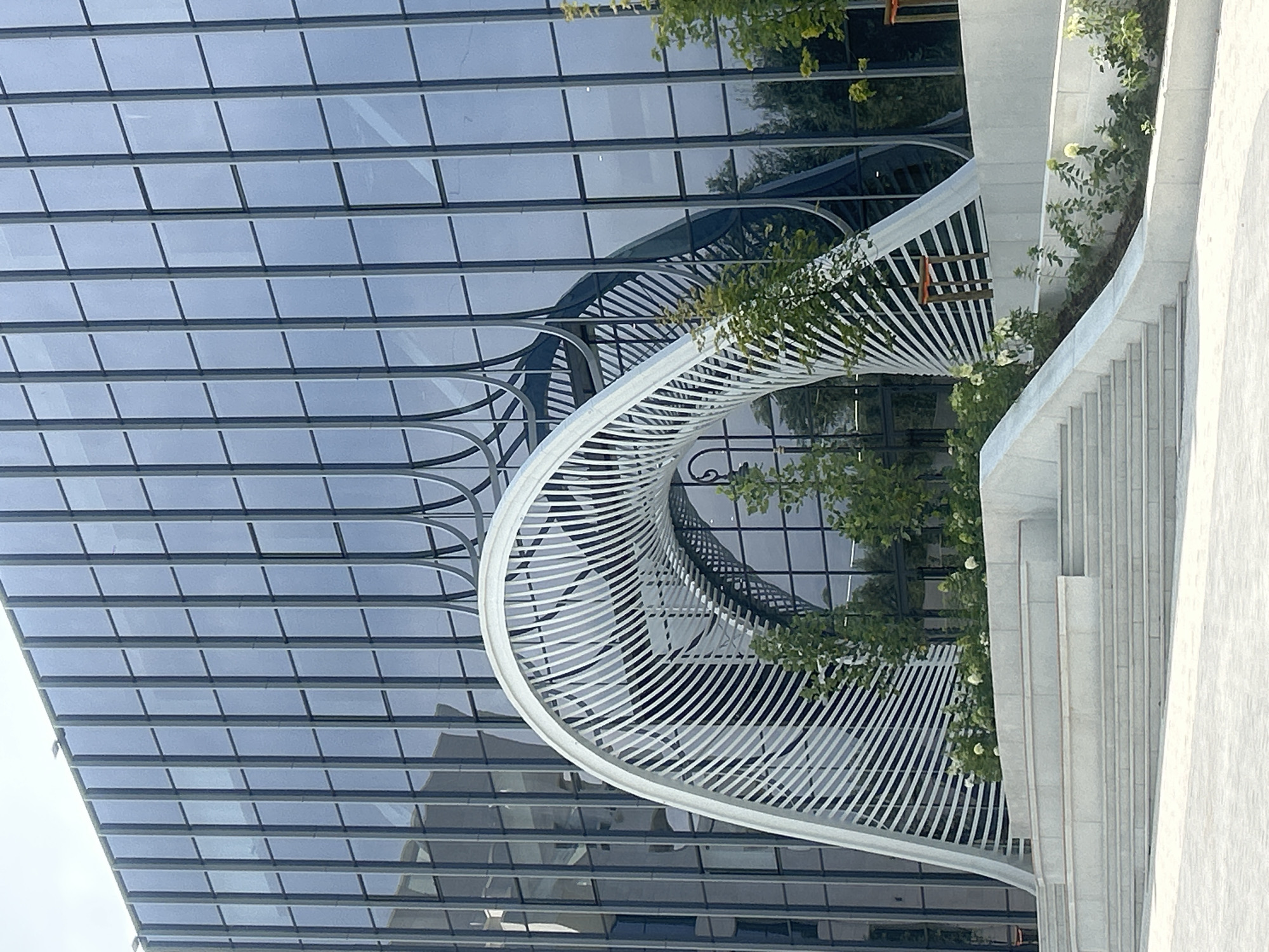 The image shows the modern architectural facade of a building in Almaty, Kazakhstan. The building features a large, reflective glass exterior with vertical mullions. In front of the building, a contemporary, white, arched metal sculpture is prominently displayed. The sculpture has a ribbed, open design and frames a reflection of the building's interior. Below the sculpture, there is a multi-tiered, light-colored stone staircase leading up to a planted area with lush green foliage and white flowers. The scene appears to be an exterior view of a public space or office building entrance.