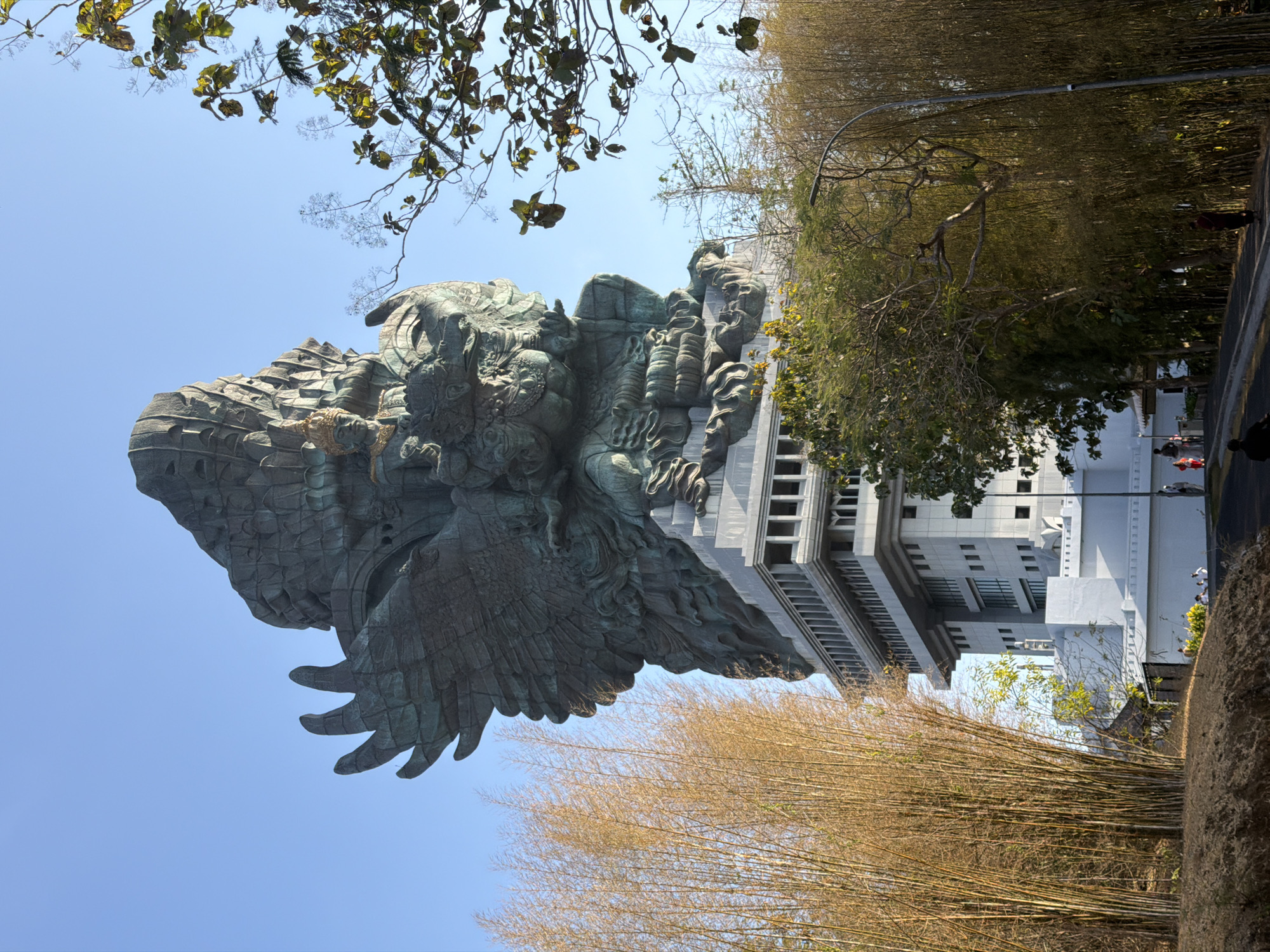 The image shows the Garuda Wisnu Kencana (GWK) Cultural Park in Ungasan, Kuta Selatan, Indonesia. The colossal statue of Garuda Wisnu, a Hindu deity, dominates the frame. It is made of bronze and is perched atop a multi-story building. The statue depicts Garuda, a mythical bird, carrying Lord Vishnu. In the foreground, there is a paved road with a few people walking and a motorcycle. To the left and right of the statue, there are dense stands of bamboo trees. The sky is clear and blue.