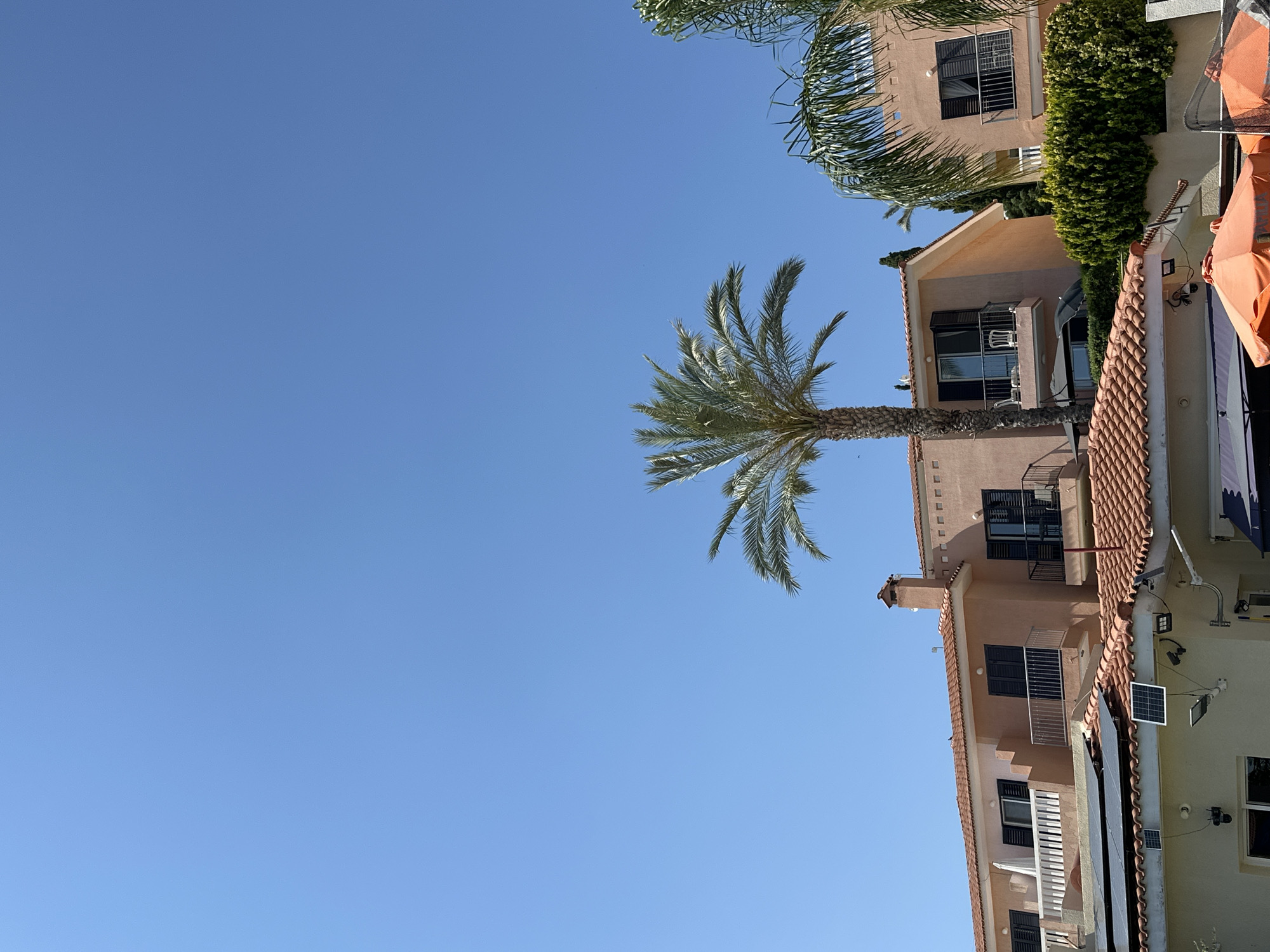 The image captures a clear, bright sky over a building complex in Peyia, Cyprus. The architecture features peach-colored facades, terracotta tiled roofs, and dark shutters on the windows. Several palm trees, characteristic of the Mediterranean climate, are visible, with their fronds reaching towards the blue sky. Some parts of the buildings show evidence of modern additions, such as solar panels and security cameras mounted on the rooftops. The scene suggests a residential or possibly a resort area, given the presence of balconies and umbrellas, hinting at leisure activities. The overall impression is one of a sunny, warm day in a coastal town.