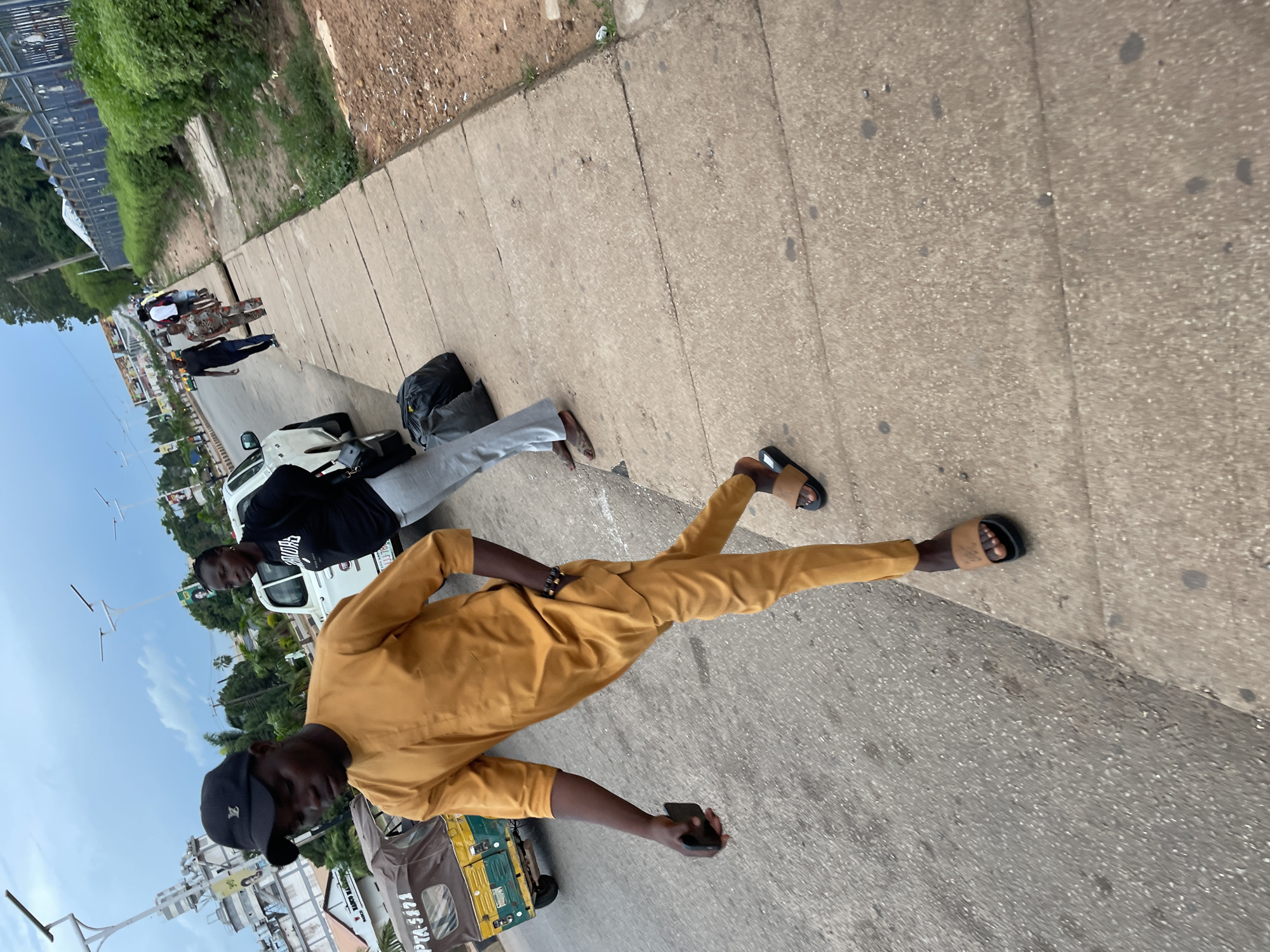 In Jos, Nigeria, on a cloudy day, a man in a mustard-colored tunic and pants walks on a sidewalk next to a street. He wears a black baseball cap and brown sandals and carries a black smartphone in his left hand. A few feet behind him, another person in a black shirt and gray pants walks, carrying two black duffel bags. A white SUV is parked on the street, and a three-wheeled vehicle is visible on the left side of the image. Buildings and trees line the street, and a blue metal fence with decorative elements is visible in the background.