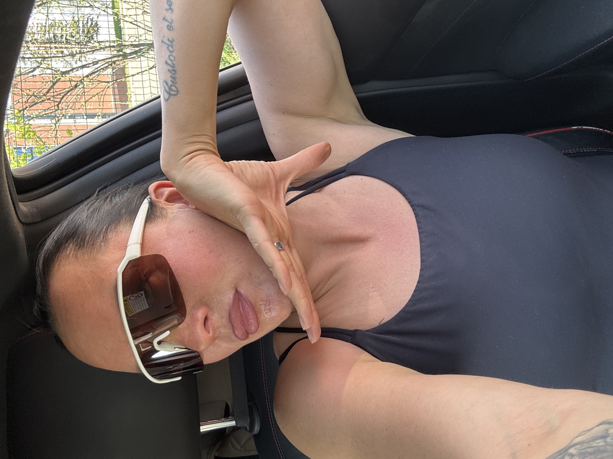 A woman is sitting in the backseat of a car, wearing sunglasses and a black tank top. Her left arm is raised with her hand near her face. A tattoo is visible on her arm. The car's interior is dark. Outside, there is a partial view of a building and trees. The location is not identifiable as Прикамский, Russia.