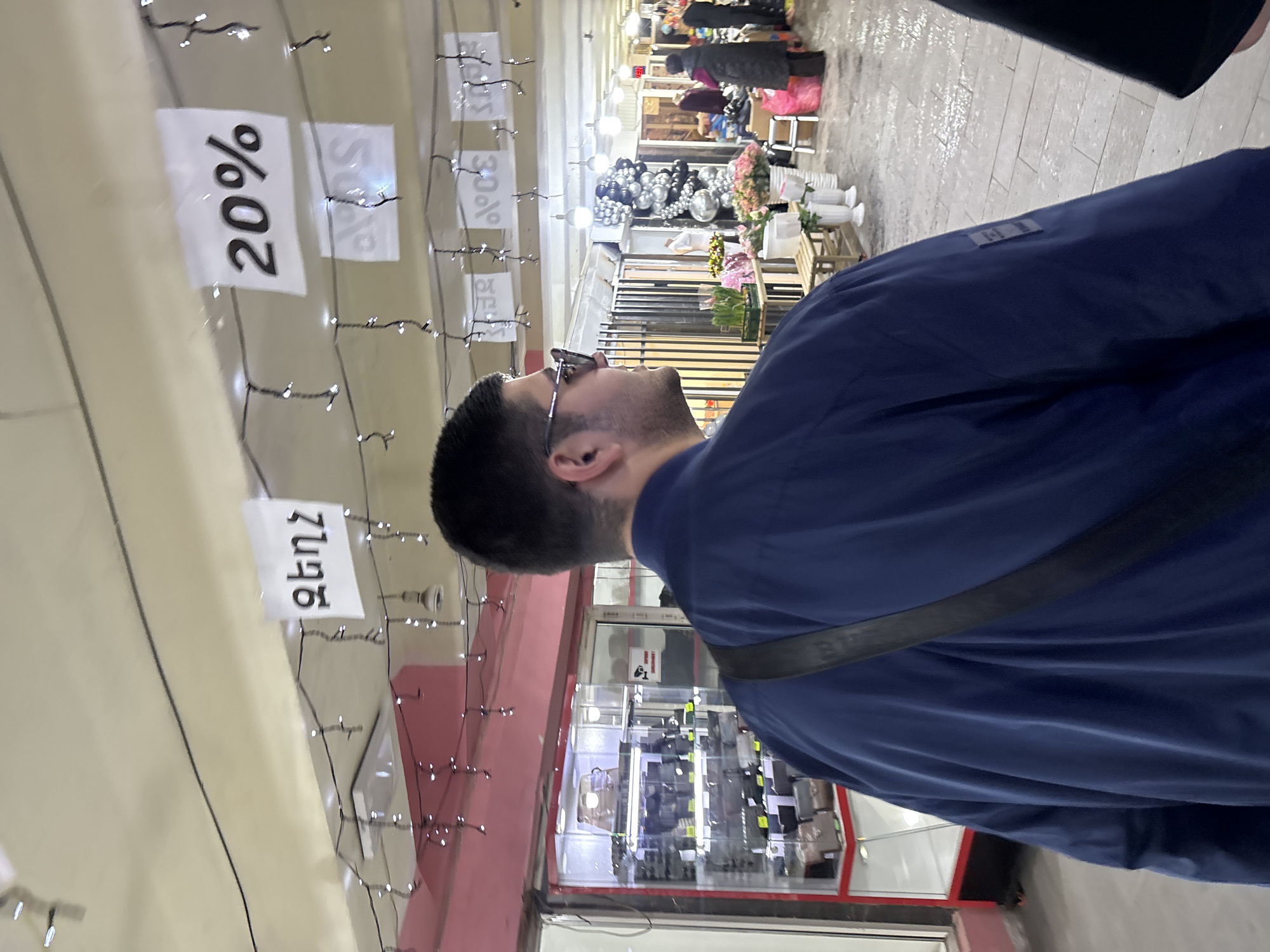 A man stands inside a shopping center in Yerevan, Armenia. He is looking up at signs hanging from the ceiling that advertise various discounts. The signs say "20%" and "30%". In the background, there are several people walking through the shopping center, as well as various vendors selling flowers and other goods. The man is wearing glasses and a blue jacket, and a black strap is visible across his back. He has short, dark hair. The shopping center has bright lighting and is lined with various storefronts.