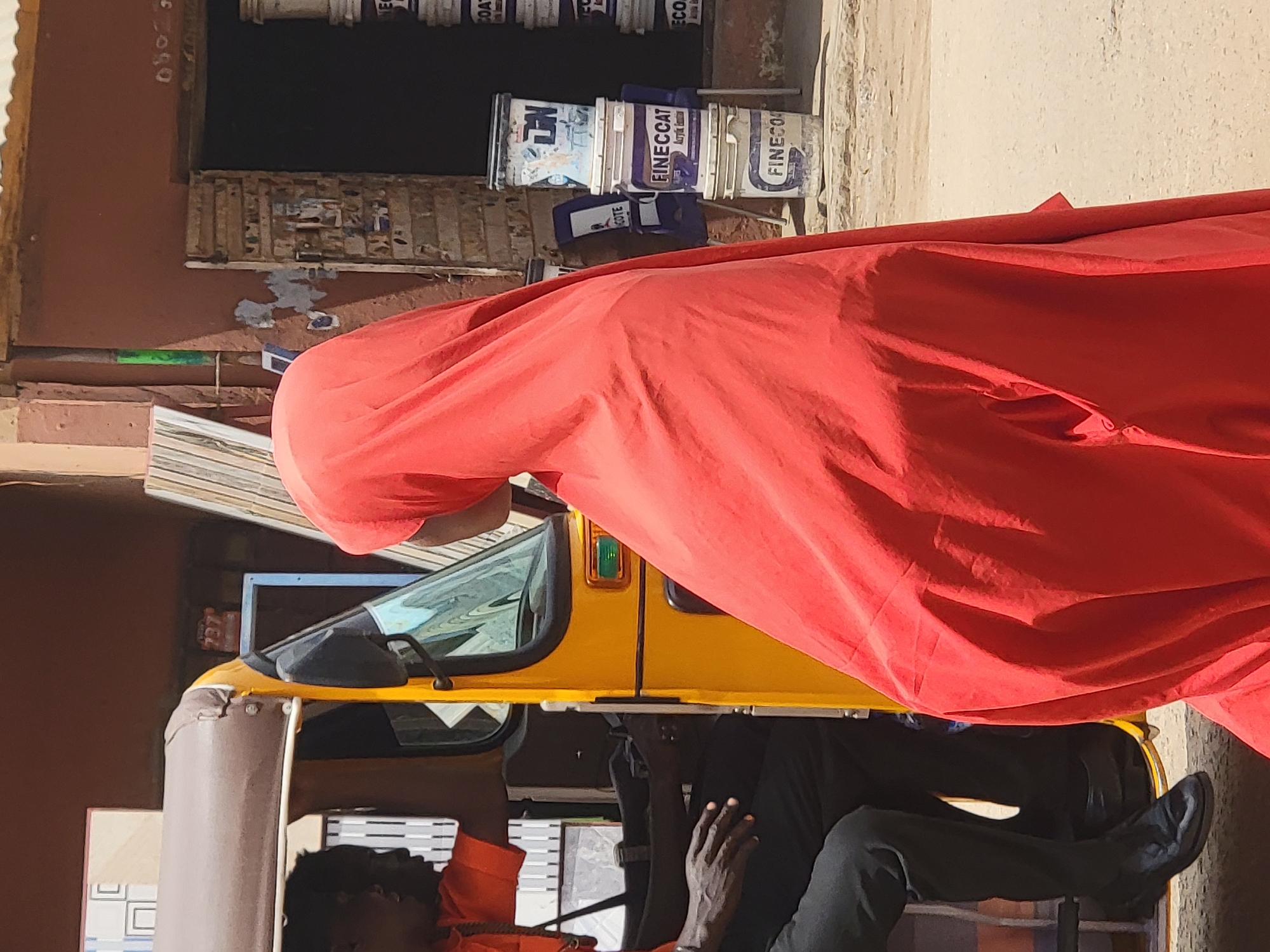 The image depicts a scene in Maiburiji, Nigeria. A yellow auto-rickshaw, also known as a "Keke," is parked on a dirt street. A man in a dark shirt and trousers is sitting in the driver's seat. The Keke is covered with a large, bright red cloth. The environment appears to be a busy street or marketplace. The background includes a building, metal pails, and other objects that suggest a commercial area.