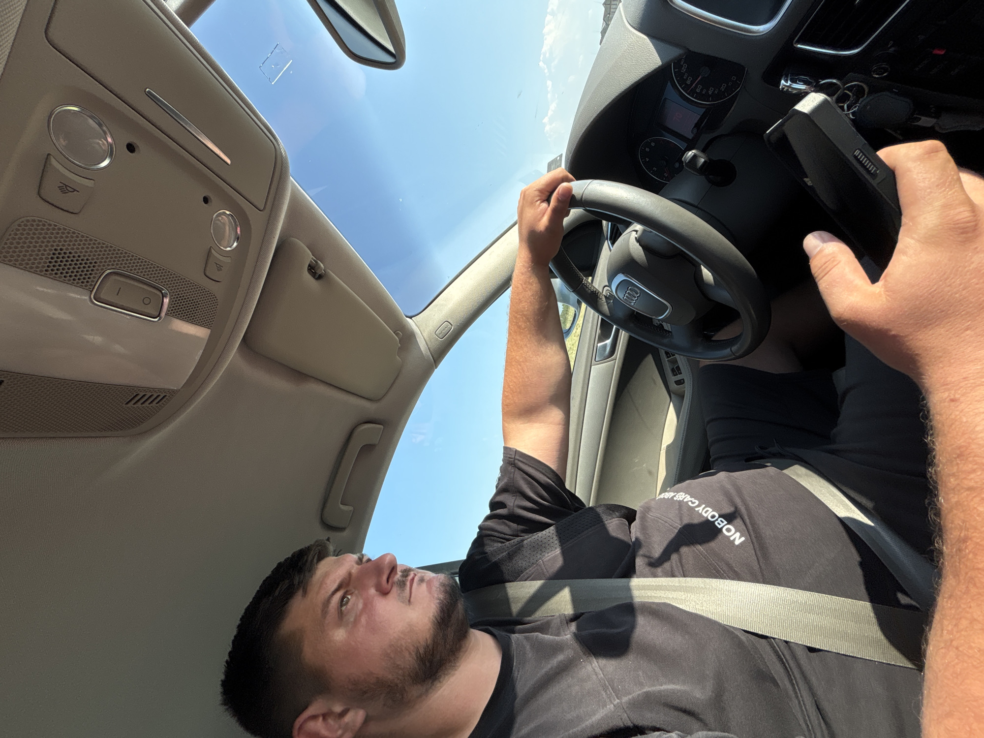 A male individual is observed driving an Audi vehicle on a clear, sunny day in Stryama, Bulgaria. The driver, appearing to be an adult male with short dark hair and a beard, is wearing a black short-sleeved t-shirt with partially visible white text that reads "NOBODY CARES" across the torso. He is secured by a light-colored seatbelt. His right hand is gripping the steering wheel, identifiable by the Audi four-ring logo, at approximately the 2 o'clock position. His left hand is holding a black smartphone horizontally, with the screen facing away from the camera, revealing a USB-C charging port and what appears to be a speaker grille on the bottom edge. The vehicle's dashboard and instrument cluster are visible, displaying a speedometer, tachometer, and a digital readout showing "70". The overhead console features interior lighting controls, a microphone grille, and other buttons. Through the windshield, a vast expanse of clear blue sky is visible, with distant landscape indicating an open environment. A rearview mirror and a sun visor are also present in the vehicle's interior.