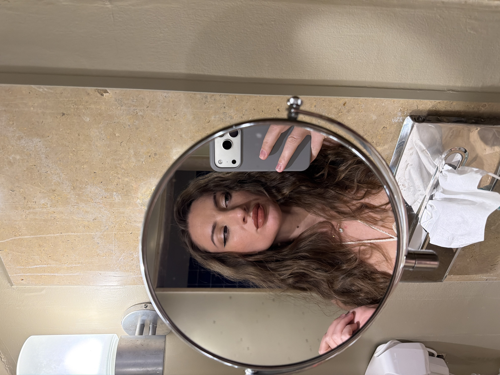 An individual is taking a selfie in a bathroom mirror. The individual is a woman with long, wavy brown hair and is wearing makeup, including eyeliner and lipstick. She is looking to her right, away from the camera. In her right hand, she holds a smartphone with a gray case, which is capturing the reflection. The bathroom has tiled walls with a beige and brown marble pattern. A vanity light fixture with a white frosted glass shade is visible on the left. Below the mirror, a chrome dispenser with white paper products is mounted on the wall. The scene appears to be in a hotel bathroom.