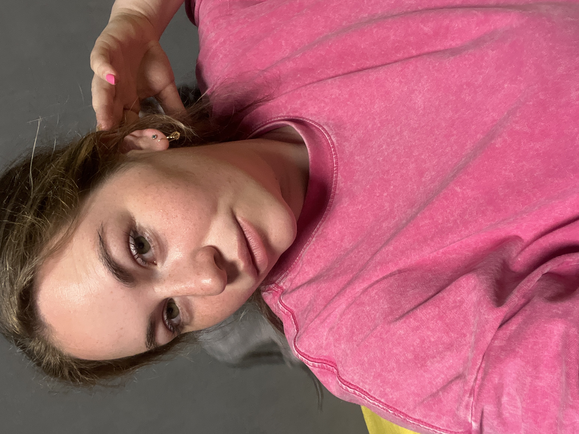 A young woman with fair skin, light brown hair, and green eyes is positioned in a selfie. She is wearing a pink t-shirt and has a piercing in her left ear. Her left hand is raised near her hair. The background is a gray or dark floor. The image does not provide enough information to identify the location as Chaikovsky, Russia.