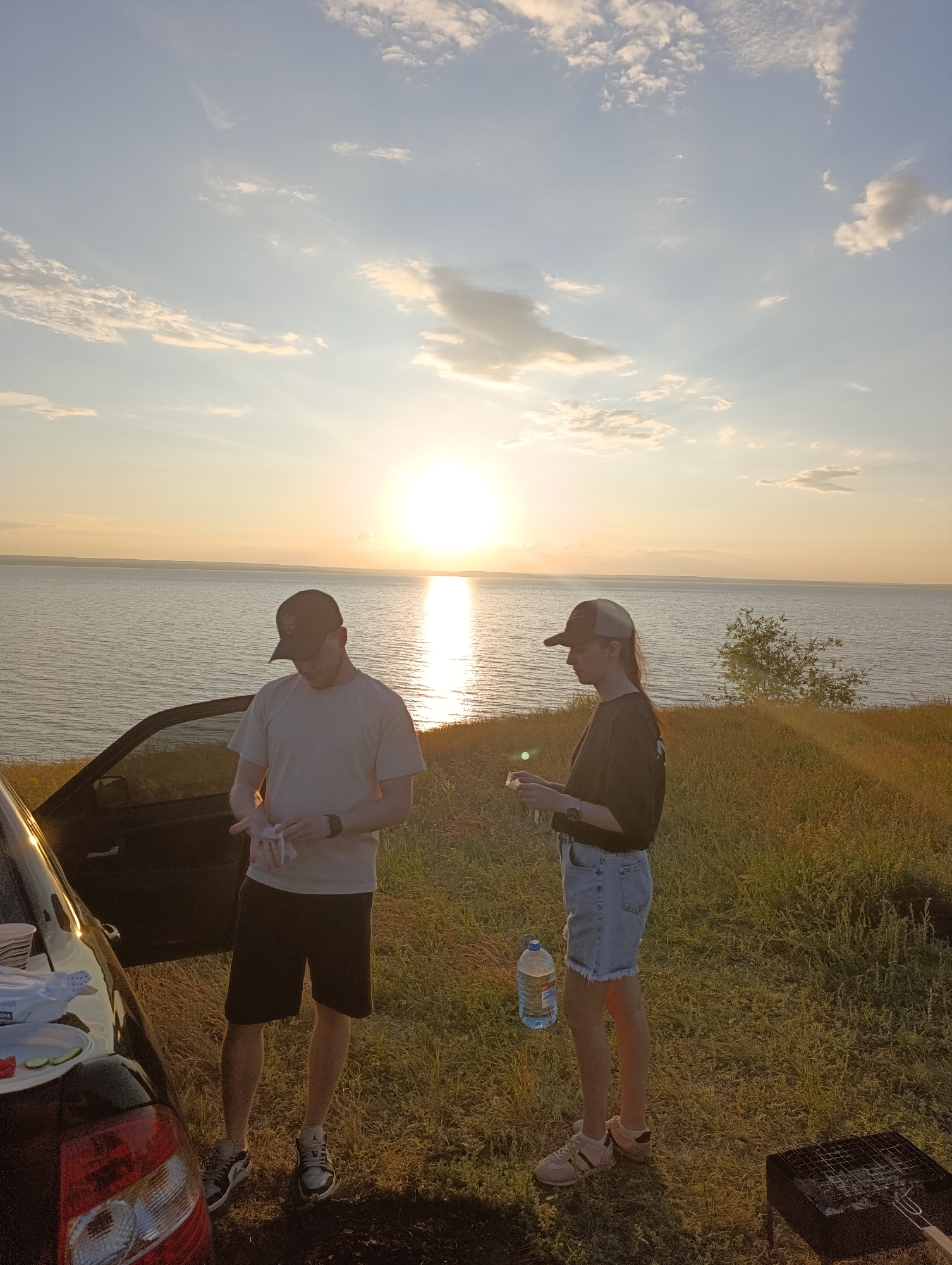 At sunset, two individuals, a man and a woman, stand on a grassy hill overlooking a wide body of water. The man, wearing a t-shirt, shorts, and a baseball cap, is holding a white cloth. The woman, dressed in a t-shirt, denim shorts, and a baseball cap, holds a large bottle of water. A car door is open to the left of the man, with picnic items visible on its surface. A portable grill is positioned on the grass to the right of the woman. The sky is filled with soft clouds, and the sun is low on the horizon, casting a warm glow over the scene. The environment appears to be a natural landscape by the water.
