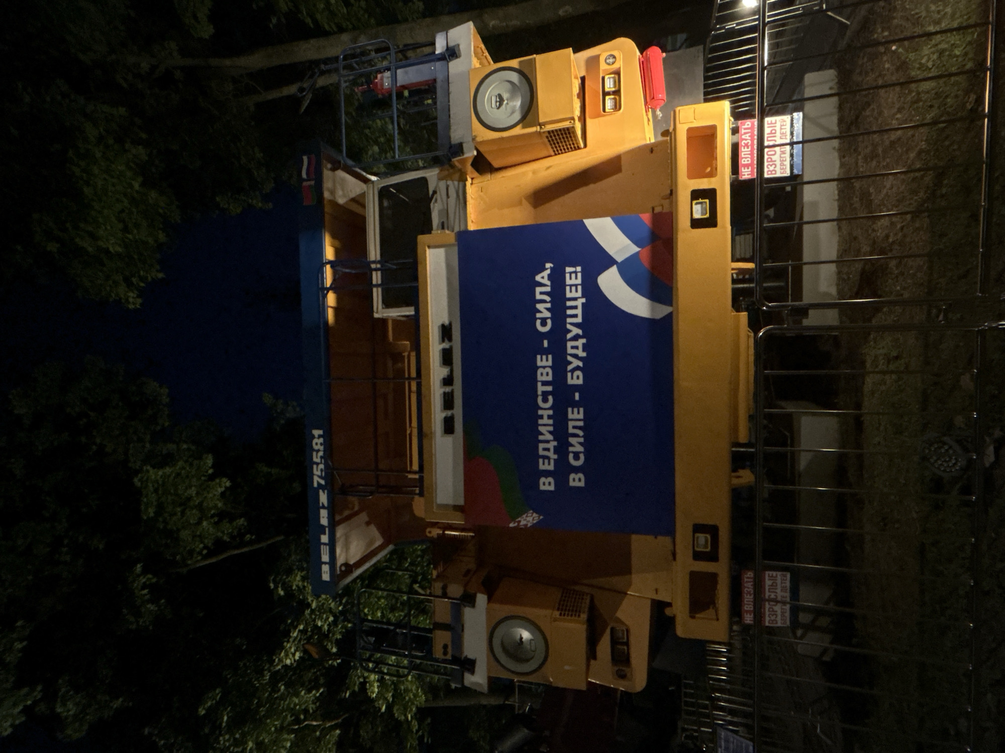The image shows a large, yellow BELAZ 75581 mining truck parked at night in a dimly lit outdoor area, likely a park or exhibition space, with trees in the background. A large blue banner with white Russian text and images of the Russian flag is displayed on the side of the truck. The text on the banner reads "В ЕДИНСТВЕ - СИЛА, В СИЛЕ - БУДУЩЕЕ!" which translates to "In unity - strength, in strength - the future!". Below the banner, two signs are attached to a metal fence. The upper sign, in red text, reads "НЕ ВЛЕЗАТЬ" (Do not enter). The lower sign, in white text on a blue background, reads "ВЗРОСЛЫЕ БЕРЕГИТЕ ДЕТЕЙ" (Adults, protect children). The overall scene suggests a public event or display. There are no people visible in the image.