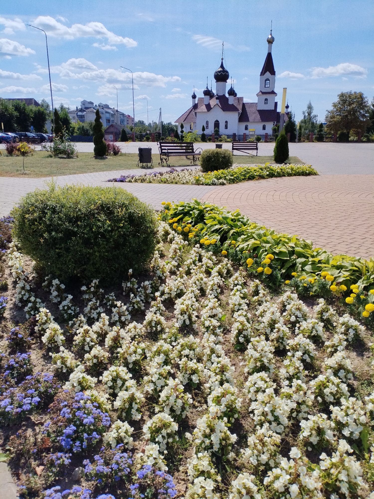 The image depicts an outdoor scene in the city of Bialynichy, Belarus, featuring a white church with multiple domes and spires in the background. In the foreground, there is a landscaped area with a variety of flowers, including white and purple varieties, arranged around a large green shrub. A paved walkway made of red bricks curves through the scene, and a park bench is visible on the left side of the walkway. The sky is blue with scattered white clouds. The overall setting appears to be a park or a public space adjacent to the church.