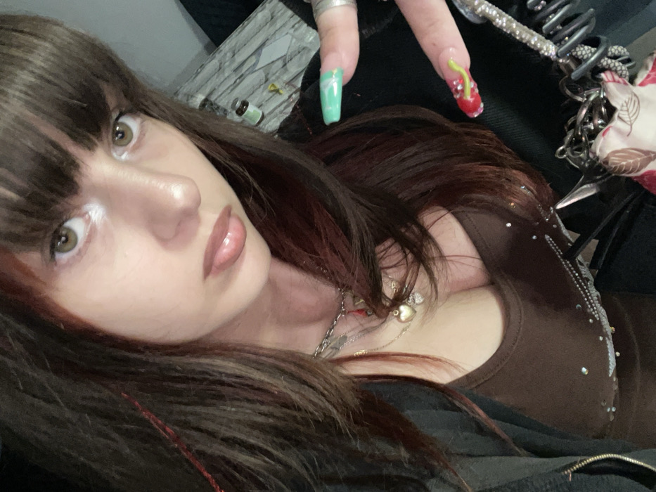 A woman with brunette hair and bangs looks at the camera. Her skin is fair, with visible highlighter on her cheeks and a glossy lip. She wears a brown tank top with rhinestone embellishments and layers of necklaces. Her nails are decorated, one green and the other red with a cherry design. The background is cluttered with various items, including a grey patterned surface, a small bottle, and accessories with chains.