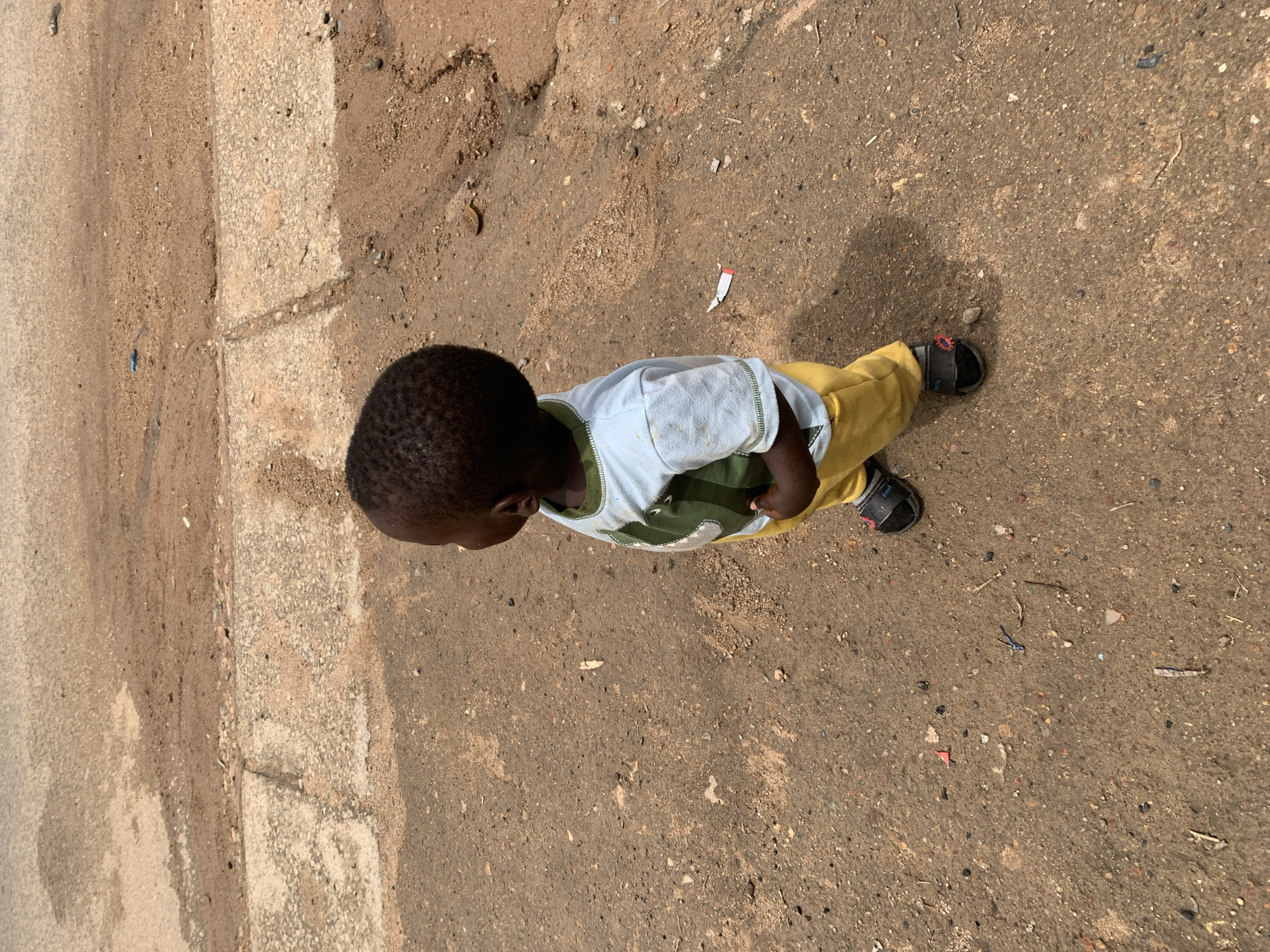 A high-angle, overhead shot captures a young child standing or walking on a dirt and gravel path in Jos, Nigeria. The child, with dark skin and short dark hair, is seen from the top of their head to their feet, looking downwards. They are dressed in a light-colored t-shirt with a green collar/trim and a subtle green pattern on the front, paired with bright yellow pants. Black sandals with small red accents, possibly floral or star-shaped, are on their feet; one red accent on the sandal has an indistinct logo or text.
The ground beneath the child is a textured mix of light brown packed earth, loose gravel, and scattered small stones. To the left, a distinct strip of lighter, coarser concrete or pavement runs vertically, separating the dirt path from another section of possibly smoother, darker ground. The overall environment appears dry and dusty. Numerous small pieces of debris are visible on the ground, including a distinctive white wrapper with red markings near the child's front-right foot, a blue plastic fragment, and various other tiny scraps of paper and plastic. The bright, even lighting suggests it is daytime with clear weather. The image focuses on the child's presence within this simple, outdoor setting, highlighting the ground and the child's apparel.