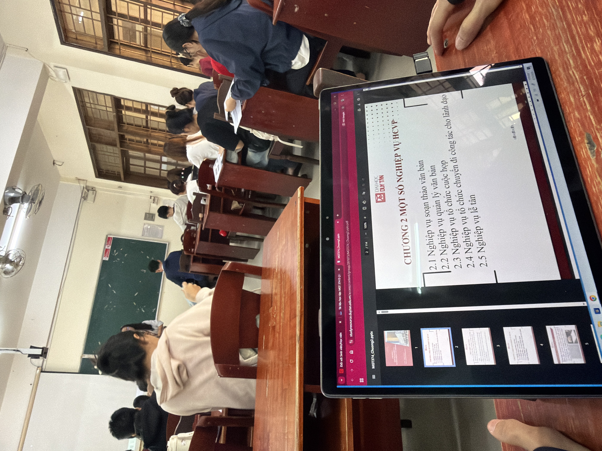 The image depicts a classroom scene at Duy Tan University in Đà Nẵng, Vietnam. Several students are visible, mostly seated at wooden desks in rows, some facing a blackboard at the front of the room (implied direction), while others are seen from behind.
In the foreground, a laptop is open on a wooden desk, displaying a presentation. A person's hand is visible next to the laptop. The screen shows a slide titled "CHƯƠNG 2 MỘT SỐ NGHIỆP VỤ HCVP" (Chapter 2: Some Office Administration Operations). Sub-points listed include "Nghiệp vụ soạn thảo văn bản" (Document drafting operations), "Nghiệp vụ quản lý văn bản" (Document management operations), "Nghiệp vụ tổ chức cuộc họp" (Meeting organization operations), "Nghiệp vụ tổ chức chuyến đi công tác cho lãnh đạo" (Organizing business trips for leaders), and "Nghiệp vụ lễ tân" (Reception operations). The Duy Tan University logo is prominently displayed on the presentation slide.
Visible browser tabs on the laptop screen confirm the academic context, showing text like "Tài liệu học tập: MGT 374 D" (Study material: MGT 374 D) and a URL pointing to "studyresource.duytan.edu.vn". The presentation is identified as "MGT374_Chuong2.pptx" and is on page 2 of 114.
The classroom environment includes a green chalkboard on the far wall, ceiling fans, and large overhead windows with patterned grilles, suggesting a daytime setting. The students appear engaged in an academic activity, possibly a lecture or a self-study session.