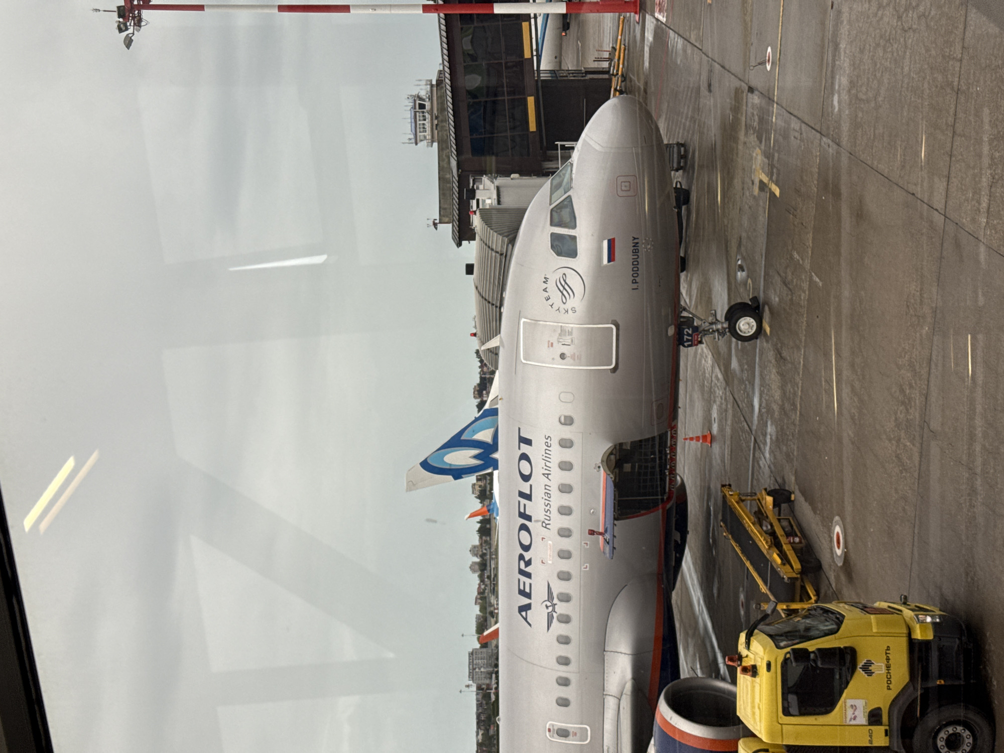The image shows an Aeroflot Russian Airlines Airbus A320 aircraft parked at an airport gate. The plane, bearing the name "I. Poddubny" near the cockpit, is connected to a mobile boarding ramp, and a yellow airport service vehicle is parked nearby. The plane's cargo hold appears to be open, indicating loading or unloading activity. The airport is likely in Sochi, Russia given it is Aeroflot and most of them exist in Russia. The backdrop features buildings and overcast sky. The image is taken from inside the airport terminal, looking out onto the tarmac.