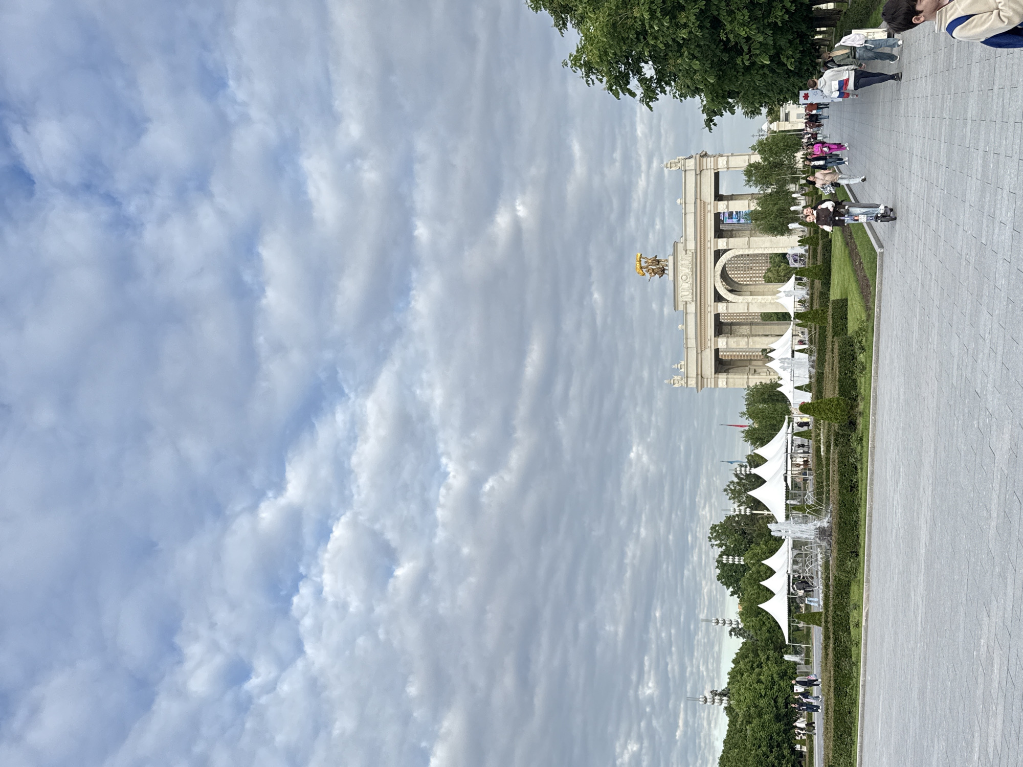 This image depicts an expansive outdoor scene, likely within a large park or exhibition complex, given the architectural style and open space, consistent with locations found in Moscow, Russia. The perspective is taken from a slightly elevated vantage point, looking along a broad, paved pedestrian pathway.
A prominent feature in the mid-ground is a grand archway, constructed from light-colored stone or concrete with classical architectural elements, including columns and intricate detailing. Perched atop the arch is a notable golden sculpture, possibly a quadriga or similar chariot with figures, which glints under the diffused light. This architectural style and the golden sculpture are characteristic of the VDNKh (Exhibition of Achievements of National Economy) in Moscow.
The sky above is largely covered with an array of scattered, textured clouds, indicating an overcast but bright daytime, likely late morning or afternoon. Lush green trees line the pathway and frame the architectural structures, contributing to a well-landscaped environment. Manicured hedges and lawns are visible alongside the main thoroughfare.
Numerous people are present, engaged in various activities typical of a public park or event. Many are walking along the paved path, some are standing in groups, and a few can be seen seated in the distance. In the immediate foreground on the right, part of a person wearing a white and blue jacket is visible. Further down the path, an individual on a scooter is discernible.
To the left of the main pathway, several distinctive white, peaked tent structures are set up. Some of these tents are situated near a fountain with visible water features, suggesting areas for refreshment, vendors, or temporary exhibits. The overall atmosphere conveys a sense of a busy but leisurely public space. No explicit text is clearly legible on any signs or structures within the visible parts of the image.