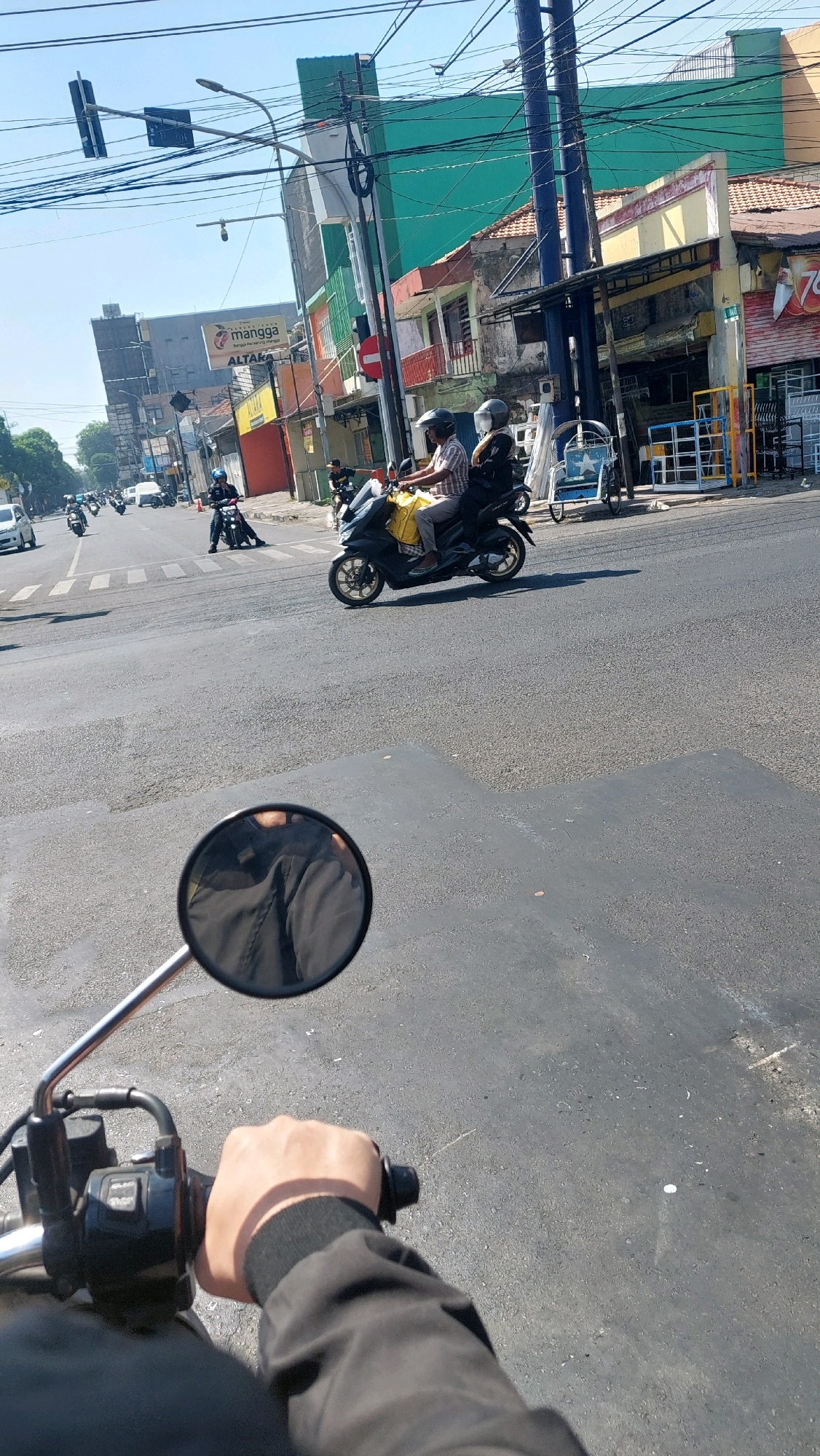 A first-person perspective captures a street scene in Sidoarjo, Indonesia, on a clear, sunny day. The viewpoint is from a motorcycle, with a rider's hand on the handlebar and a side mirror reflecting the rider's upper body. In the middle ground, a black motorcycle carries two passengers, the one in front wearing a plaid shirt and helmet, the rear passenger in black with a helmet. A bright yellow bag is strapped to the back of this motorcycle. Traffic is present, with other motorcycles visible in the background. Buildings line the street, featuring colorful facades and awnings. Overhead power lines crisscross the sky. The road surface is asphalt, with clear lane markings. A pedestrian crossing is visible on the left side of the road. The overall impression is of a typical urban streetscape in Indonesia.