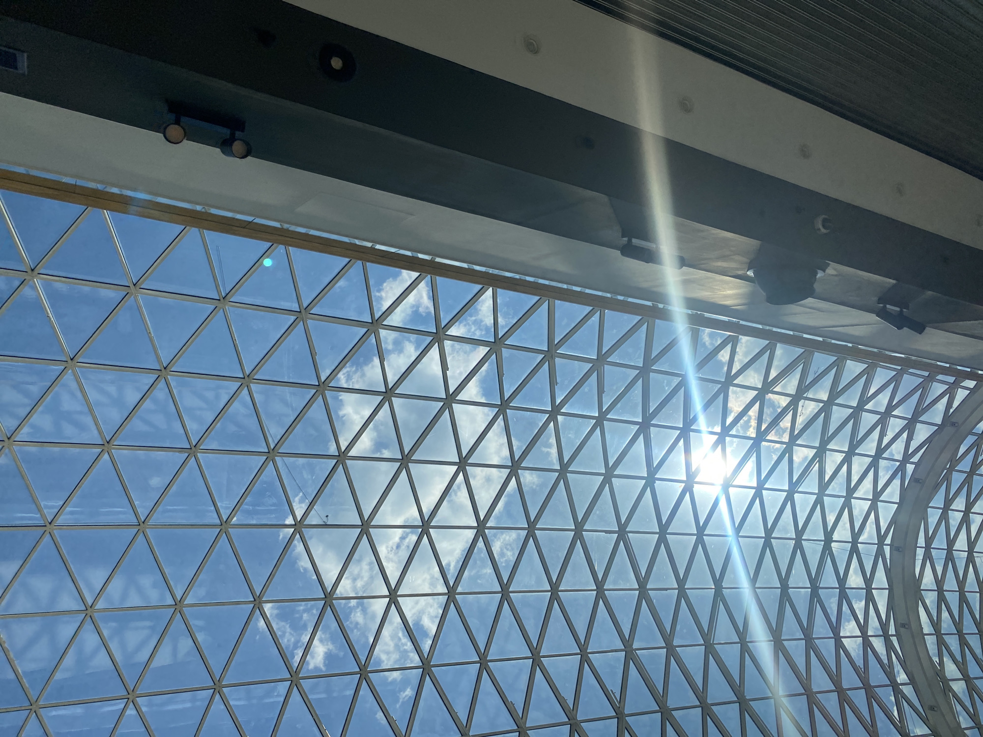 The image shows a view upwards through a triangular lattice ceiling, with a clear blue sky and scattered white clouds visible through the openings. Sunlight is shining through, creating a bright glare. Above the ceiling is a structure with lights, including spotlights and recessed lighting. The lattice pattern suggests the structure might be located in the city of Perm, Russia.