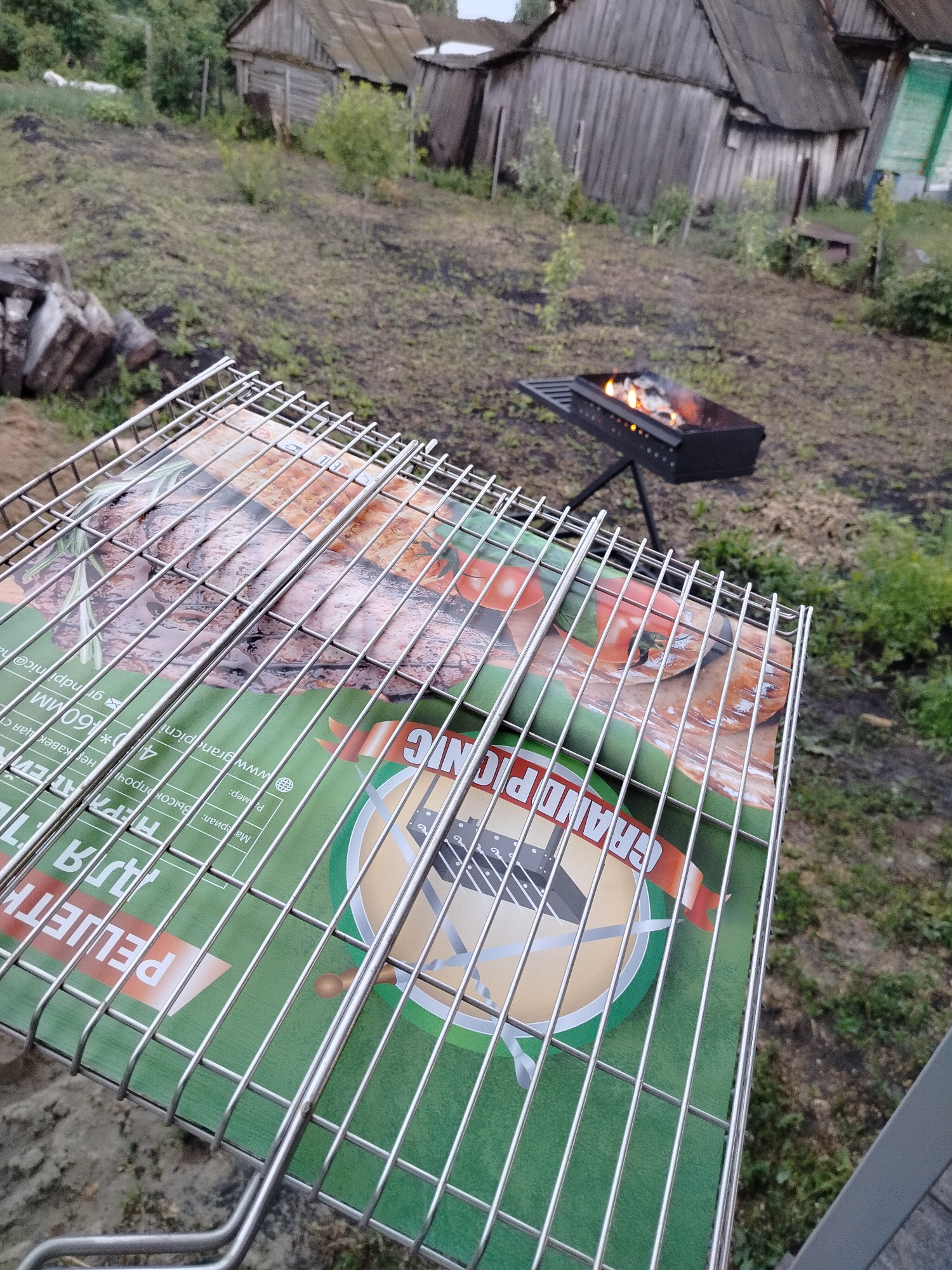 This media file depicts an outdoor scene with a focus on a grill grate placed over a green packaging with text. In the background, a traditional Russian dacha or rural setting is visible, characterized by weathered wooden buildings and unkempt vegetation. A portable charcoal grill with glowing embers is positioned on the ground.
The subjects are primarily inanimate objects: a metal grill grate, green packaging, and a barbecue grill. The scene is set in a rural or dacha area, suggesting an outdoor activity like a barbecue or picnic. The time of day appears to be late afternoon or early evening, as indicated by the soft lighting and overcast sky, suggesting possibly a cloudy day. The overall impression is one of a casual, possibly spontaneous, outdoor meal preparation.
Notable details include the text on the green packaging, which appears to be in Russian. Key phrases that can be deciphered include "GRAND PICNIC," "PELJETH," and specifications like "40*160мм" (likely dimensions) and "нержавеющая ст" (stainless steel). The phrase "Материал: Высокопрочі" might refer to the material being high-quality. The website "www.grandpicnic.ru" is also visible, confirming the brand name.
The visual composition, with the grill grate in the foreground, creates a sense of anticipation for the grilling to begin. The background elements of dacha buildings and nature contribute to the rustic and homely atmosphere. There are no people visible in the frame, emphasizing the objects and the setting.