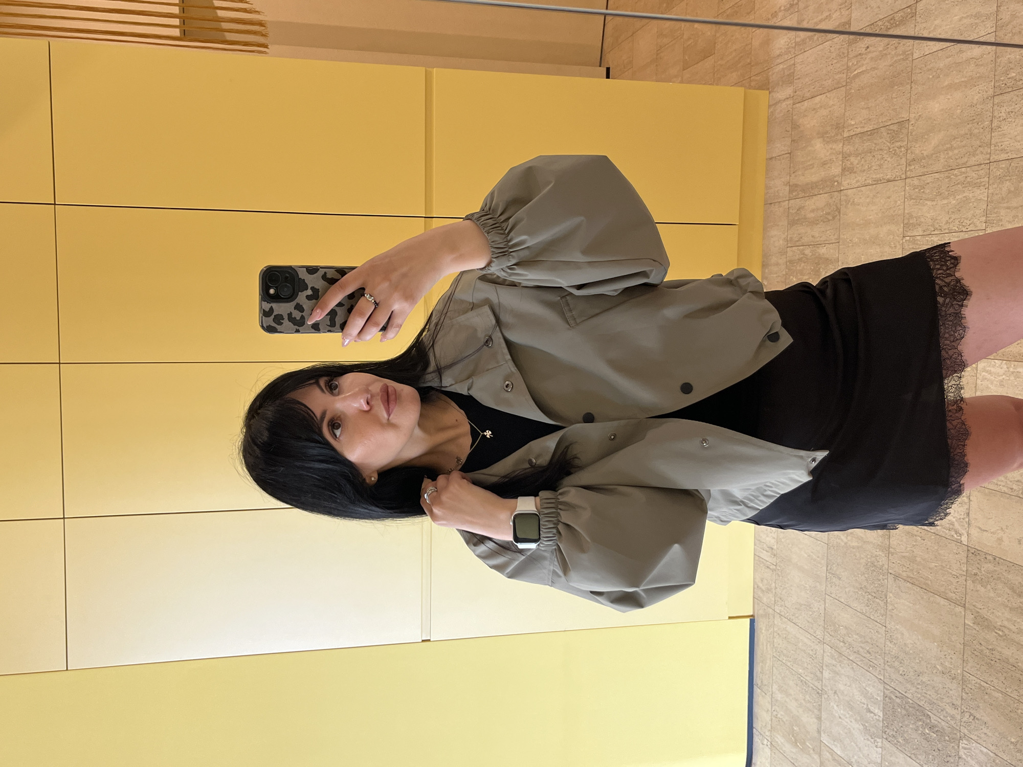 A woman is taking a mirror selfie indoors. She has dark hair with bangs and is wearing a black dress with lace trim, a light green jacket with button details, a silver necklace, and a silver watch. She is holding a phone with a patterned case. The background includes a light yellow wall with panels and a tiled floor. The photo was taken in Sirius, Russia.