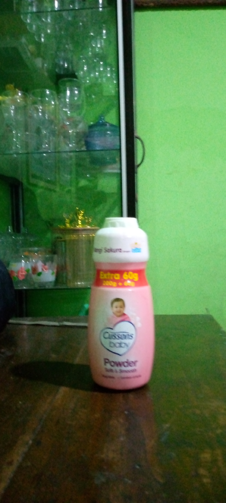 A light pink Cussons Baby powder bottle is positioned on a dark brown wooden surface. The bottle has a white cap and features a label with an image of a baby, the brand name "Cussons BABY," and the word "Powder." Above this, a yellow band reads "Extra 60g." In the background, a green wall is visible, and to the left, a glass cabinet contains various glass items, including what appear to be cups and a blue decorative object. The scene is located indoors.
