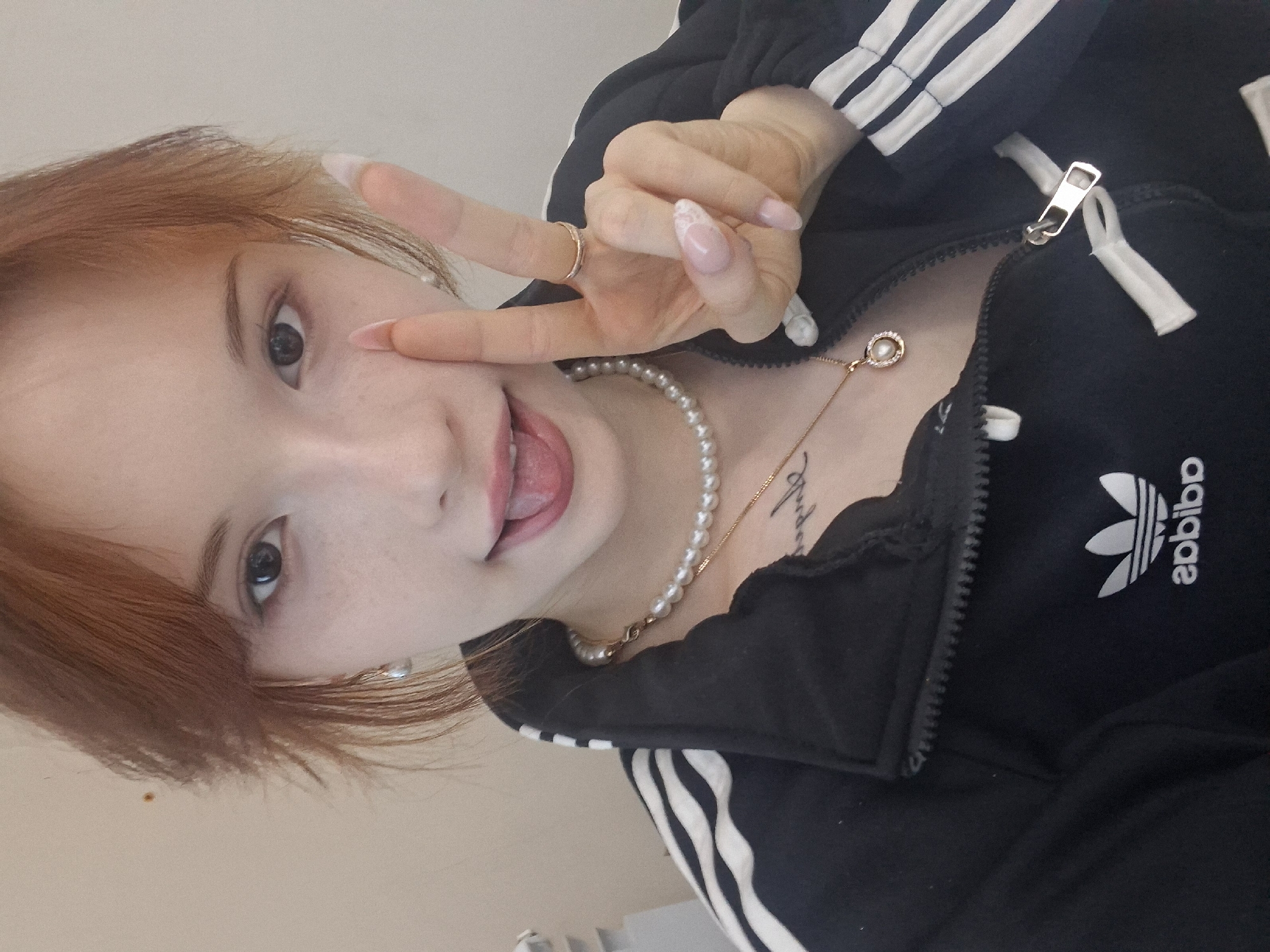 The image is a selfie taken by a young woman with reddish-brown hair and light skin, showing her face and upper body. She is wearing a black Adidas jacket with white stripes on the sleeves, a pearl necklace with a pendant, and a ring on her right hand. Her tongue is sticking out, and she is holding up two fingers in a peace sign. The background is a plain, light-colored wall. The location of this image in the city of Chaikovsky, Russia is not discernible from the provided image or context.
