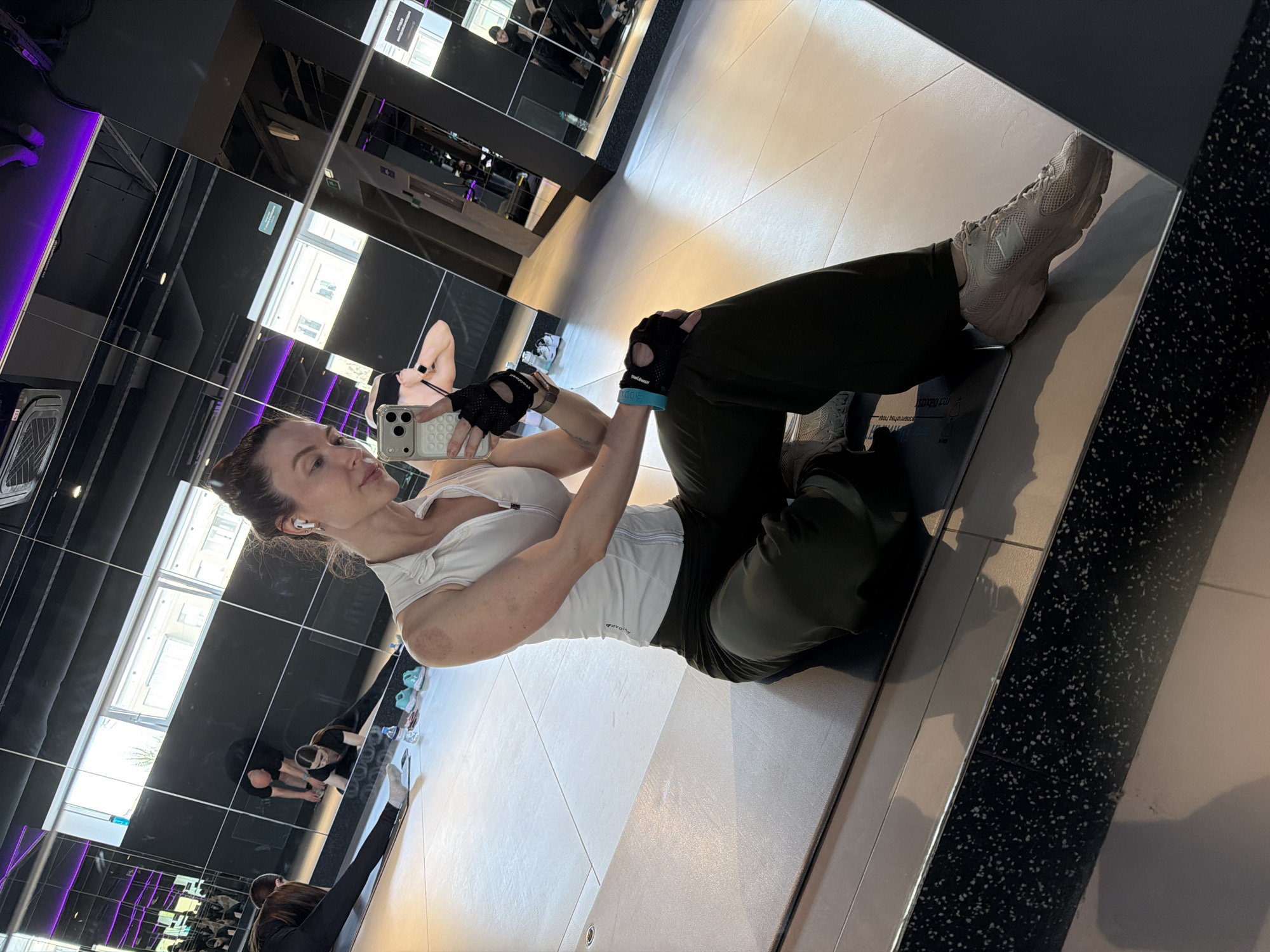 A woman lies on her back in a gym, holding a smartphone up to take a selfie. She is wearing a white athletic top, dark pants, workout gloves, and light-colored sneakers. Her hair is pulled back and she has an earpiece in her ear. The gym has mirrored walls, reflecting the woman and other people exercising in the background. The location is Perm, Russia.