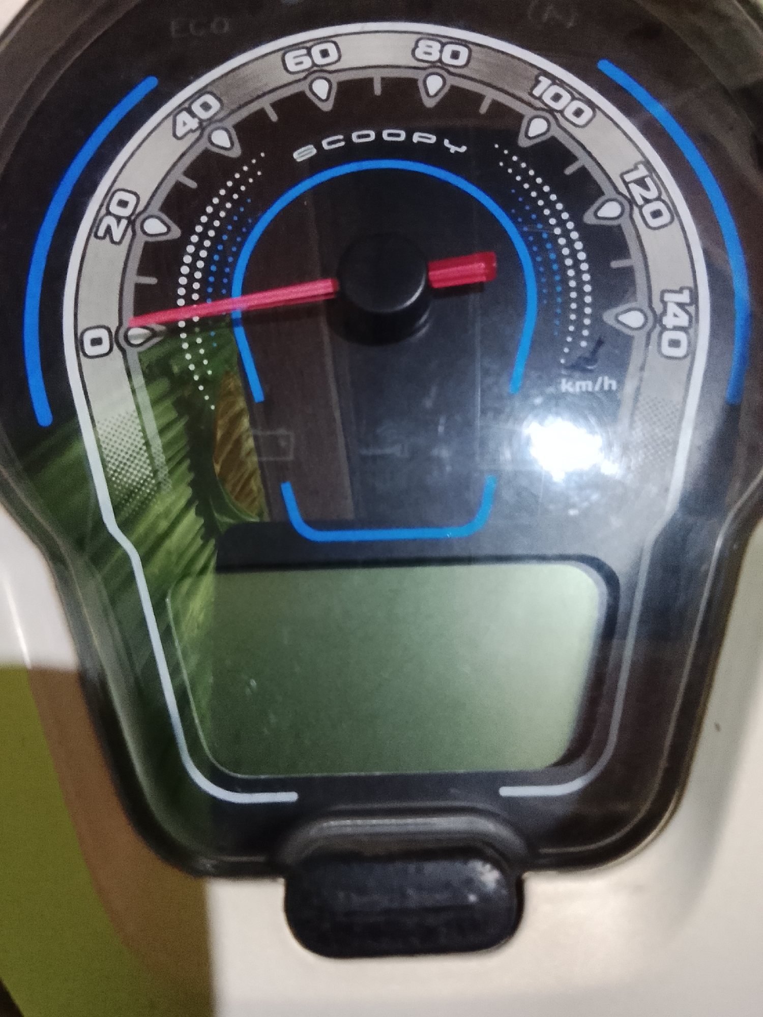 This is a close-up, high-angle shot of a motorcycle's speedometer and digital display. The speedometer dial has markings for 0, 20, 40, 60, 80, 100, 120, and 140 km/h. A red needle points just above the 0 km/h mark, indicating the motorcycle is likely stationary or moving very slowly. The word "SCOOPY" is visible above the needle. To the right of the speedometer, a green digital display is located. The background is mostly dark with some green and brown reflective elements. The location of Tanjongbabahkrueng, Indonesia is not discernible from the image.
