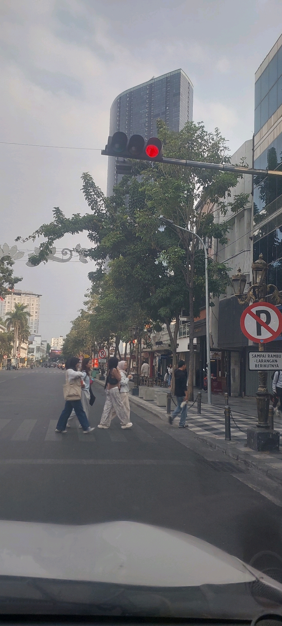 The image depicts a street scene in Surabaya, Indonesia, likely during the late afternoon or early evening based on the lighting. In the foreground, the hood of a vehicle is visible, suggesting the photo was taken from inside a car. The street has a pedestrian crossing with four individuals crossing from left to right. Two women are in the front, followed by two others. They are dressed in casual, light-colored clothing, with one woman wearing a hijab.
Further down the street, several trees line the sidewalk, which appears to be a commercial area with shops and buildings. A prominent tall, modern building with a curved facade dominates the background. A traffic light is visible, showing a red signal. A "No Parking" sign is mounted on a lamppost on the right side of the street, along with a sign written in Indonesian that translates to "Until the next traffic light." The sky is overcast.