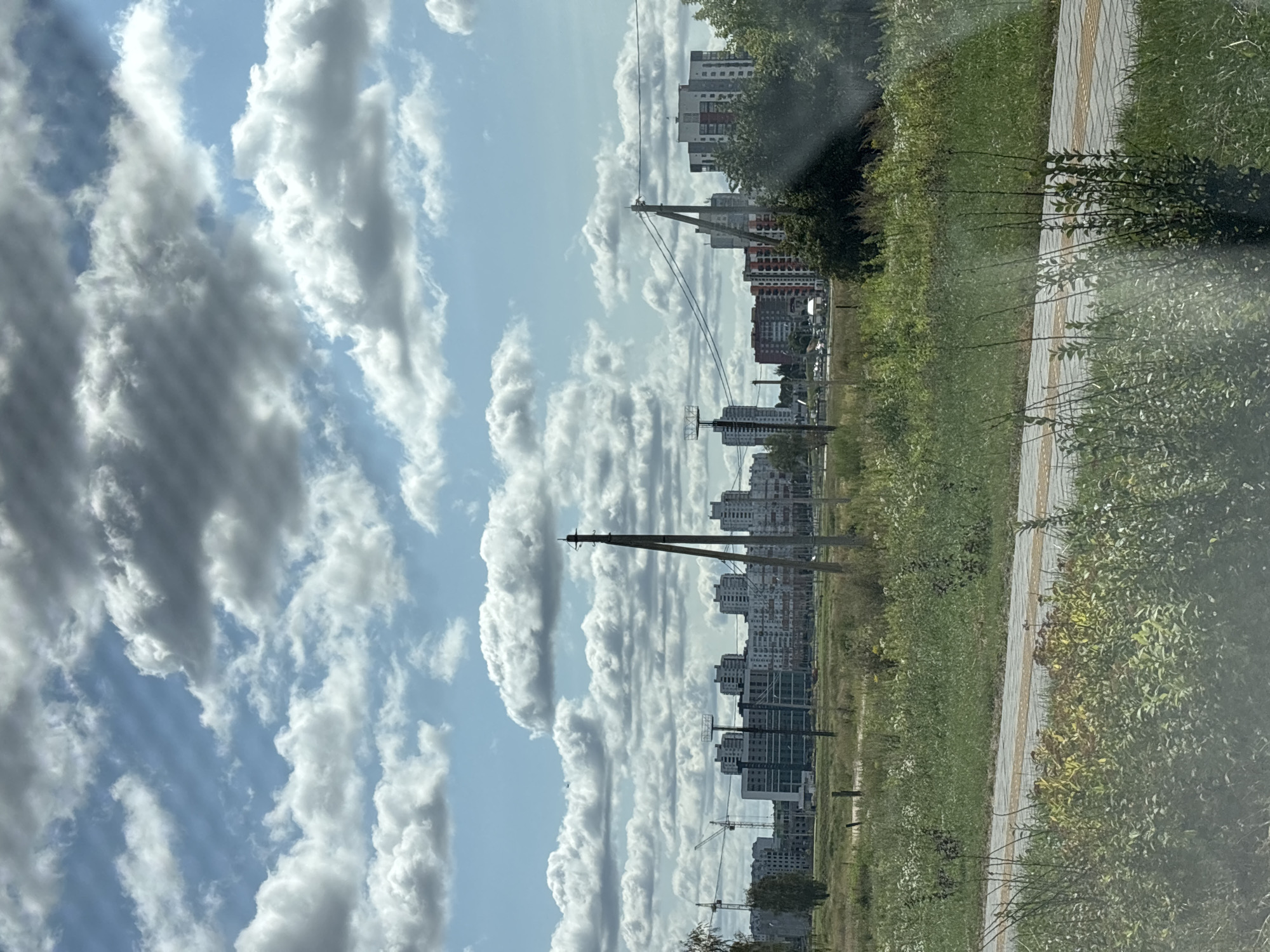 The image depicts a landscape view with a cloudy sky above a line of modern apartment buildings in the distance. In the foreground, there is a patch of green grass and other vegetation, with a paved path. Several utility poles with wires are visible in the middle ground. In the background, construction cranes suggest ongoing development in the area. The overall scene is one of urban expansion with undeveloped land surrounding it. It is possible this scene is in Minsk, Belarus, given the architectural style of the buildings and the presence of construction.
