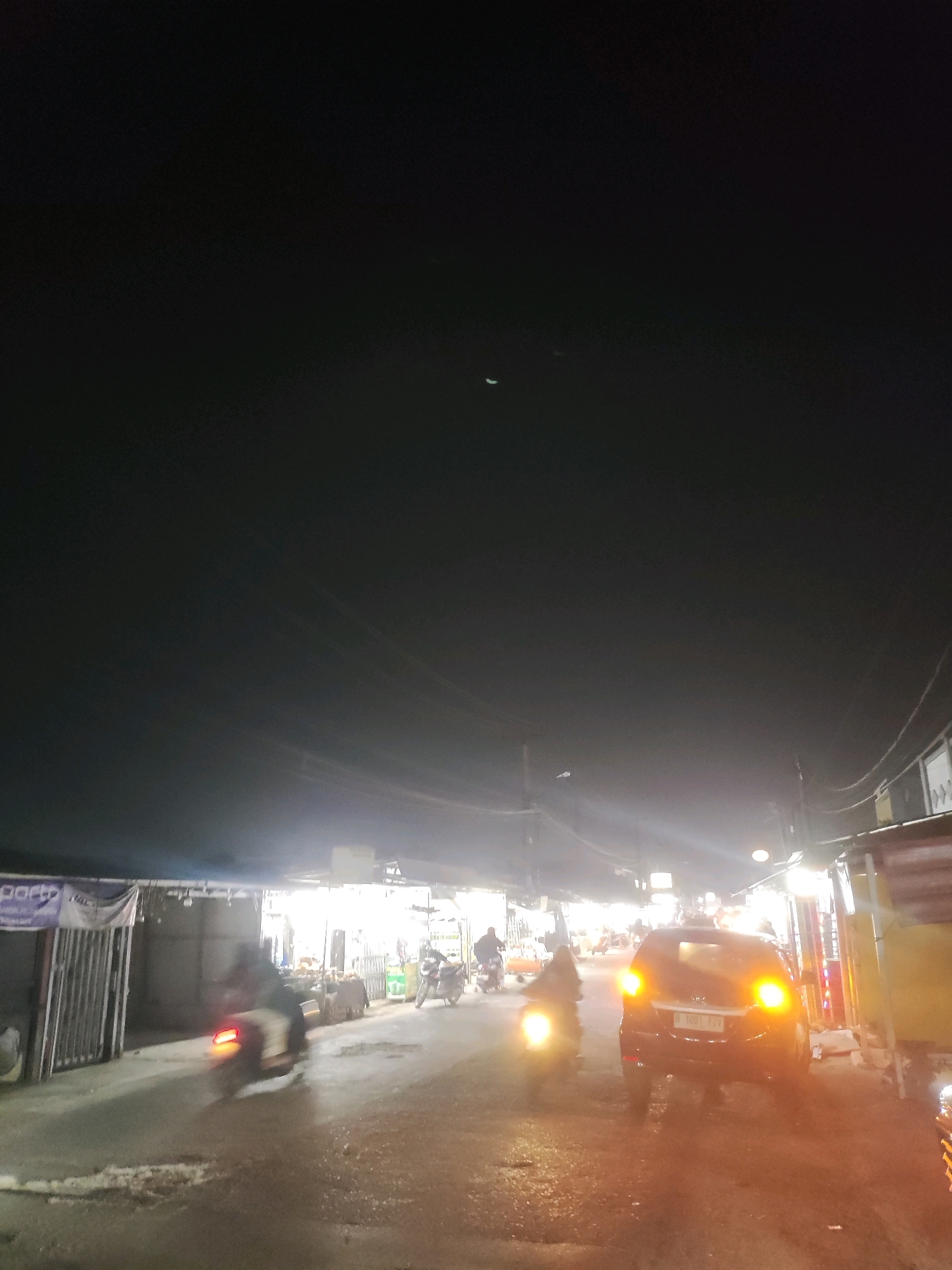The image depicts a nighttime street scene in Telagamurni, Indonesia. A dark-colored car with illuminated taillights is parked on the right side of a wet road. To the left of the car, a motorcycle is being ridden by an individual, with its headlight and taillight glowing. Further down the road, other vehicles and a few figures are visible, suggesting ongoing activity. The street is lined with buildings and stalls, many of which are illuminated by artificial lights, creating a bustling market-like atmosphere. The sky is dark, with a faint crescent moon visible.