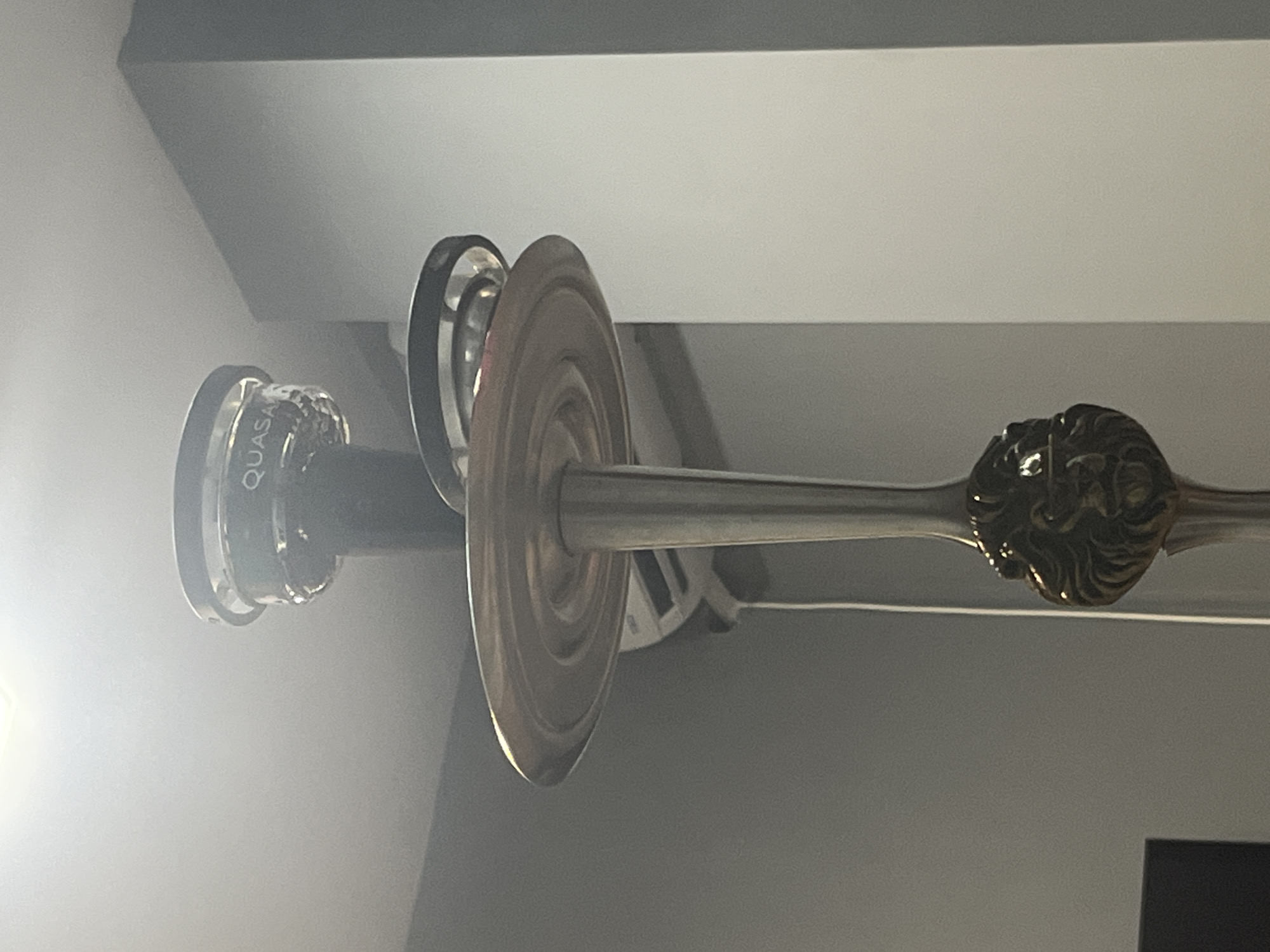 The image shows a close-up, upward view of a hookah pipe. The pipe is made of metal and features a decorative lion's head design near the base of the stem. At the top of the pipe, there is a black cylindrical component with the word "QUASAR" visible, suggesting it might be a brand name or model. Above this, there appears to be a glass component or a screen. A round, metallic tray is positioned below the black cylinder. The background consists of a light-colored ceiling with a bright light source in the upper left corner, and a gray wall. An air conditioning unit is partially visible on the wall behind the pipe. The scene appears to be indoors, in a residential or commercial setting. There is no discernible location in the city of Montana, Bulgaria, or any other specific geographical information provided within the image.