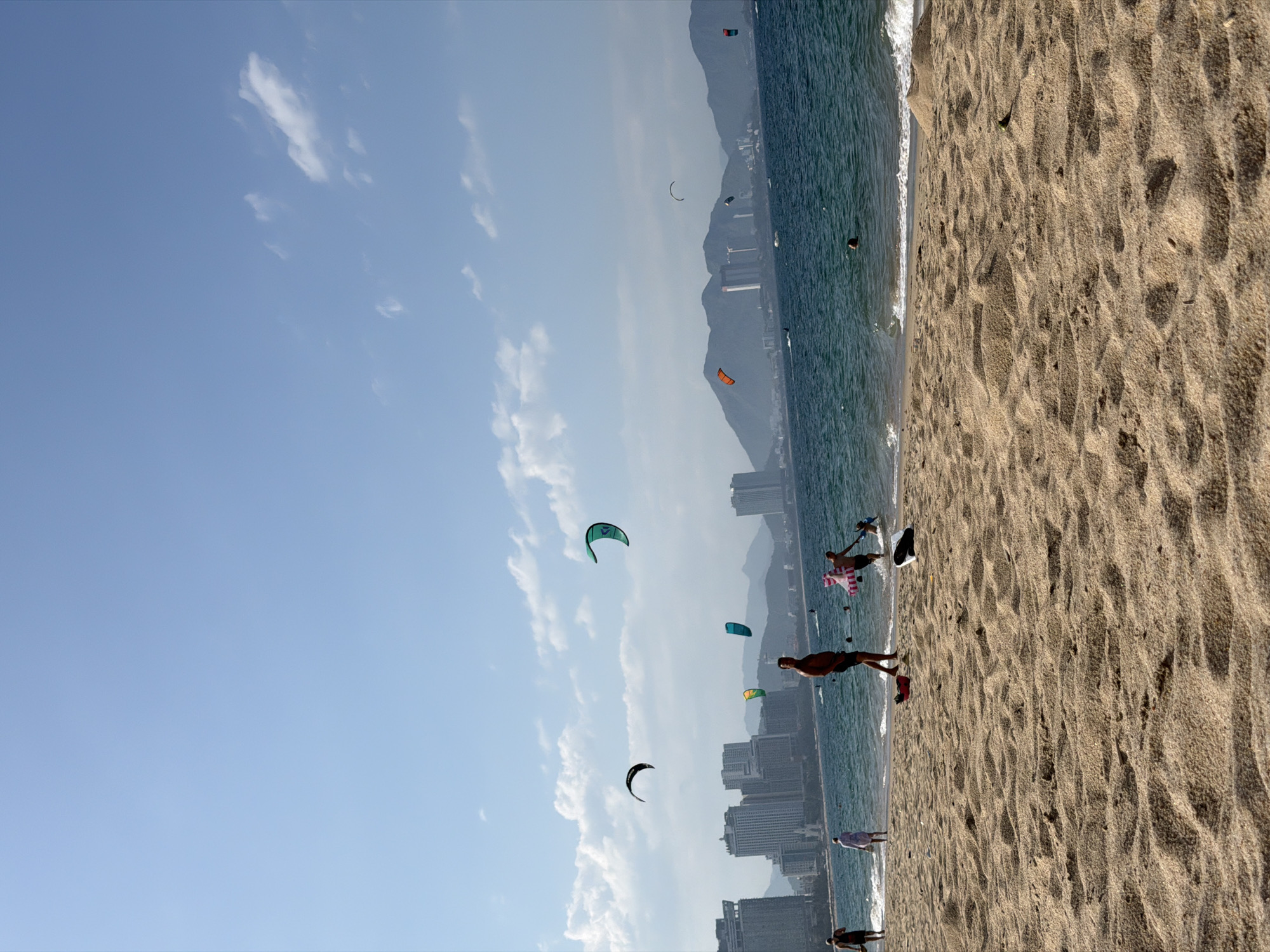 The image depicts a coastal scene in Nha Trang Ward, Vietnam, under a clear blue sky with scattered white cumulus clouds. A wide, light brown sandy beach occupies the foreground, extending to a blue-green ocean with gentle waves.
**People and Activities:** Approximately eight individuals are visible. On the far left, a person in dark swimwear walks towards the water. A second person, dressed in light-colored clothing, stands at the water's edge with their back to the viewer. Near the center, a man with a bare torso and dark shorts stands in the shallow water, facing partially left. To his right, another man in dark shorts is assisting a small child or person with a pink and white striped inflatable ring in the shallow water. Further out in the water, only the head of a swimmer is visible.
**Kitesurfing:** Several kites are airborne, indicating kitesurfing activity. A large dark-colored kite is visible in the upper left quadrant. A prominent green kite arches in the upper right quadrant. Smaller kites, including one yellow-green and one blue-green, are closer to the water's surface in the mid-ground. Distant kites, one orange-red and one dark, are also visible over the water near the mountainous horizon.
**Objects and Environment:** On the sand near the water, a red object (possibly a bag) and a white object are present. A small green object rests on the sand further in the foreground. In the distance, a city skyline composed of multiple modern high-rise buildings lines the coast, with a range of hazy, green-tinged mountains forming the backdrop. A faint wake from a powered watercraft is discernible in the far left of the ocean.
