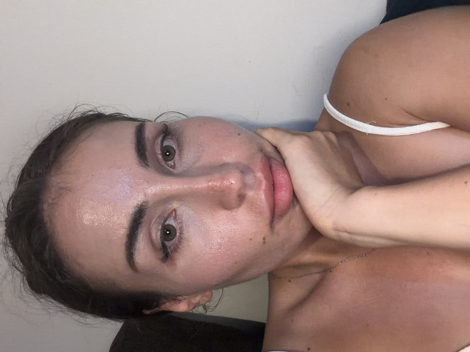 A close-up, slightly angled, medium-shot photograph shows a fair-skinned woman with dark hair pulled back from her face. She is resting her chin on her left hand, with her elbow on her lap. Her eyes are open and directed towards the camera. She is wearing a white tank top, and a thin silver necklace is visible around her neck. The background consists of a plain, light-colored wall. The lighting appears to be from the front and slightly to the side. The image does not contain any information or visual cues to suggest a location within Tersefanou, Cyprus.