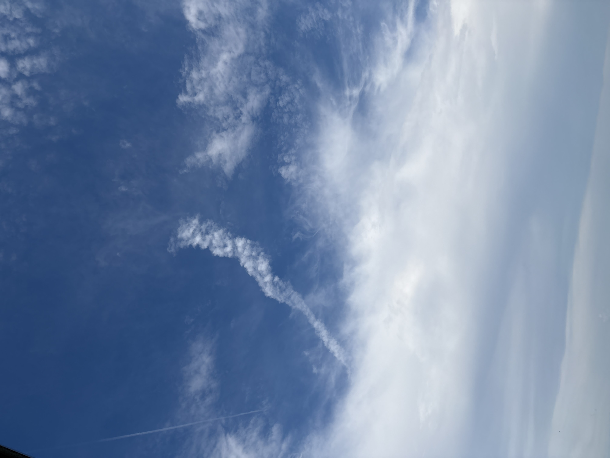 The image shows a wide-angle view of a bright blue sky with scattered white clouds. A prominent contrail from an aircraft is visible, stretching diagonally from the upper left to the center of the frame, appearing as a thin white line that then expands into a wispy, textured cloud formation. Towards the bottom of the image, the clouds become denser and more overcast, transitioning to a softer, diffused light. The scene is devoid of any human presence, structures, or identifiable landmarks that would specifically place it within Bönnigheim, Germany. The environment is exclusively sky.