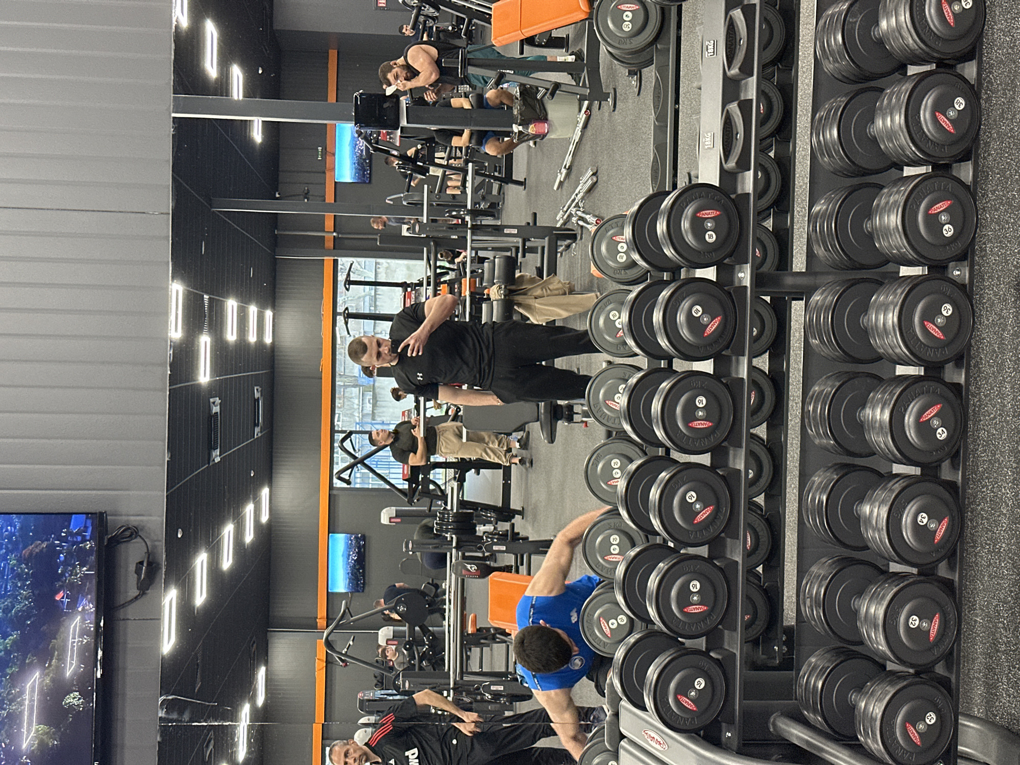 The image depicts the interior of a gym. A rack of black dumbbells, labeled "PANATTA" with varying weights (e.g., 32kg, 34kg, 16kg, 18kg), stands prominently in the foreground on the right side of the frame. People are exercising and interacting with the equipment. One man wears a blue shirt, and another a black shirt. A television is visible on the left side. The gym has orange accents and appears to be well-lit. The precise location within Plovdiv, Bulgaria is not specified.