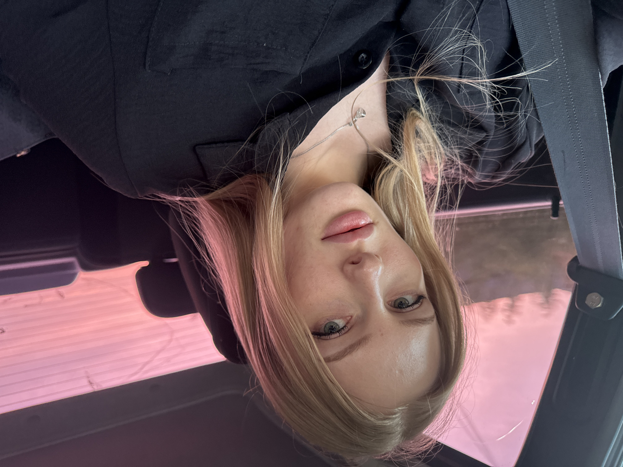 A woman with blonde hair and fair skin is wearing a black shirt and a silver necklace. She is sitting in a car with a seatbelt across her chest. The car is located in Кондратово, Russia, as visible through the pink tinted glass of the car.