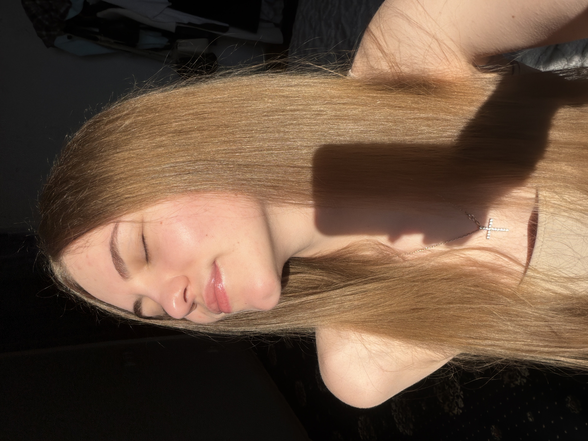 This is a close-up, horizontally oriented photograph of a young woman from the chest up, with her head tilted back and eyes closed. She has long, light brown hair that is illuminated by sunlight, creating a warm glow and casting a shadow across her hair and shoulder. Her skin appears smooth and lightly flushed, with a subtle sheen on her nose and lips, suggesting a natural, dewy look. She is wearing a light-colored garment, possibly a top or dress, and a delicate silver necklace with a cross pendant is visible. Her eyebrows are neatly shaped, and her lips are slightly parted, giving her a serene and peaceful expression. The background is dark and out of focus, with some indistinct shapes that could be furniture or clothing. The lighting suggests it is daytime, with sunlight streaming in from the side, highlighting the texture and color of her hair. The overall mood of the image is tranquil and intimate. There is no visible text in the image.