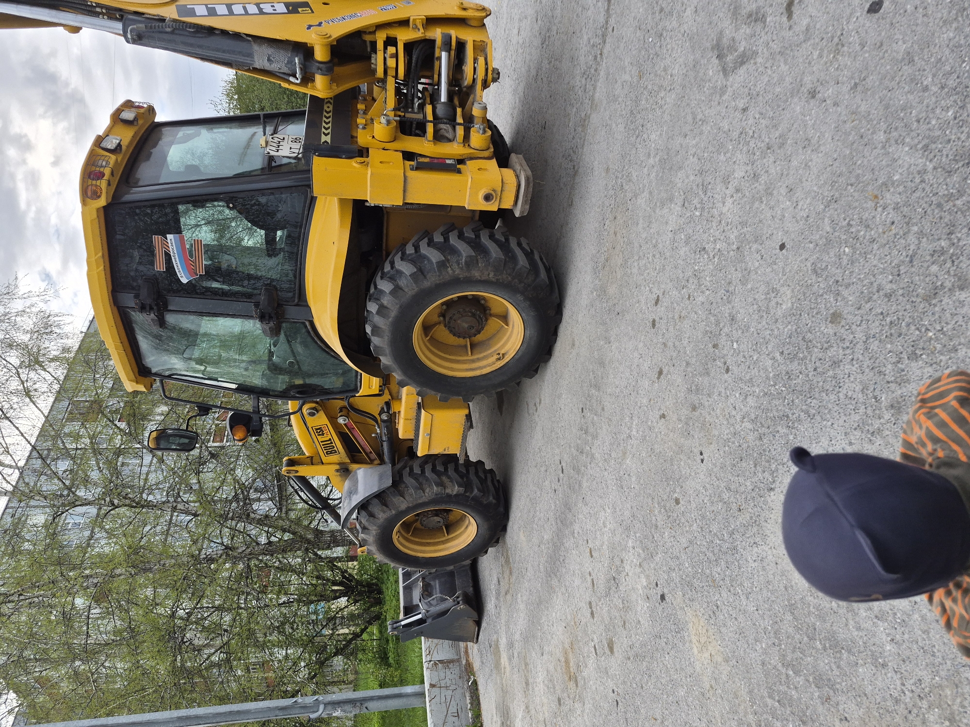 A yellow construction vehicle, a BULL 45X backhoe loader, is parked on a paved surface, likely a street or parking area. The backhoe has prominent tires and a front-end loader. The vehicle's cab features a Russian license plate with the number "4442 YT 86" and an insignia containing the letter "Z," associated with support of the Russian military. A dark blue baseball cap rests on the pavement with a portion of a striped sleeve visible. The image shows trees and a multi-story building in the background. Based on the license plate, the vehicle may be located in the Khanty-Mansi Autonomous Okrug region of Russia, but not Tobolsk. The location of Tobolsk cannot be determined from this photograph.