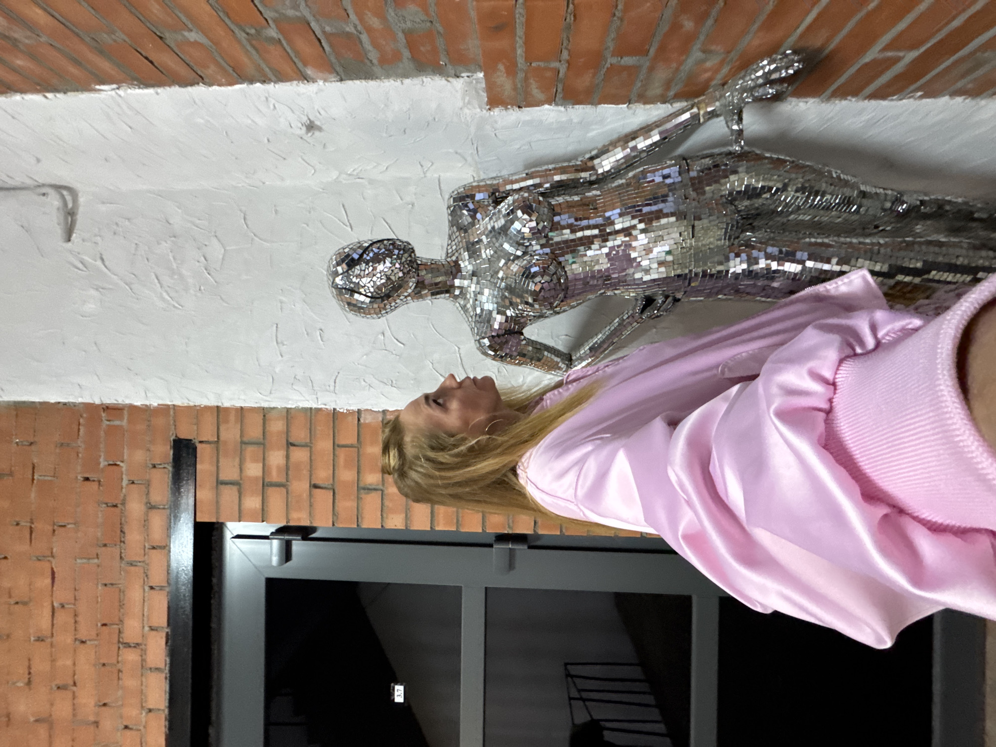 A woman in a pink satin outfit poses next to a mirror mosaic sculpture of a woman. The background includes a section of a brick wall, a white textured wall, and a window. The location is implied to be Saint Petersburg, Russia.