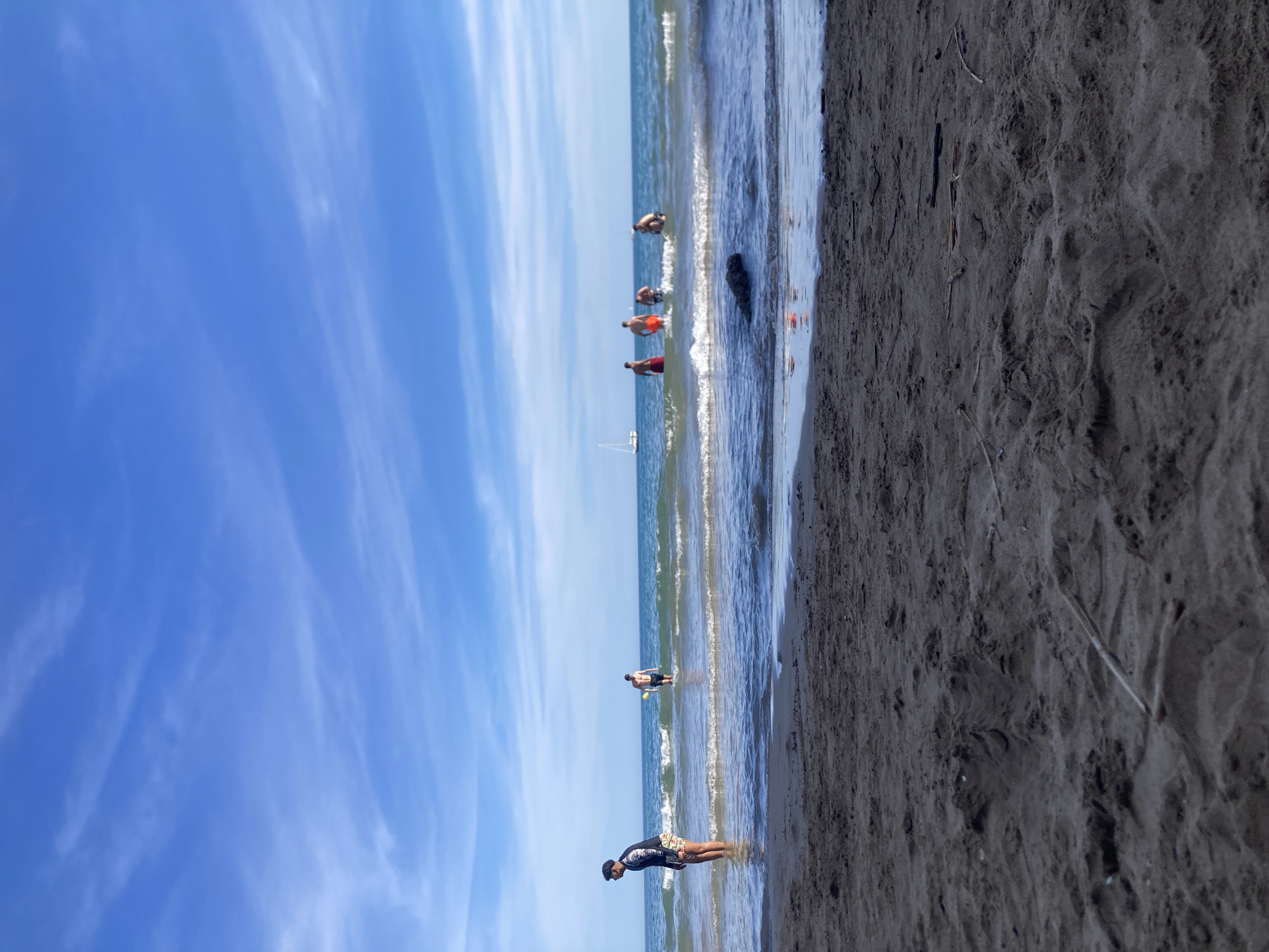 A group of people are in the water at a beach. In the foreground, dark sand is visible with some debris scattered on it. The water is blue with white waves. In the background, a sailboat is on the water under a bright blue sky with wispy white clouds.
