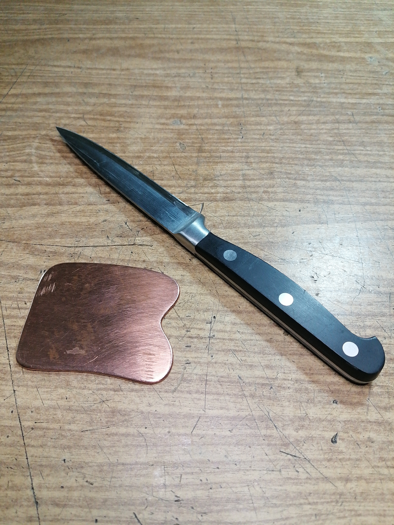 A close-up shot shows a knife and a copper tool on a wooden surface. The knife has a black handle with white rivets and a silver blade. The copper tool is heart-shaped and has a metallic sheen. The wooden surface appears to be a workbench or table, with visible grain and scratches. The lighting is natural and casts soft shadows. The scene is static and does not depict any activity or interaction. The location is not discernible from the image.
