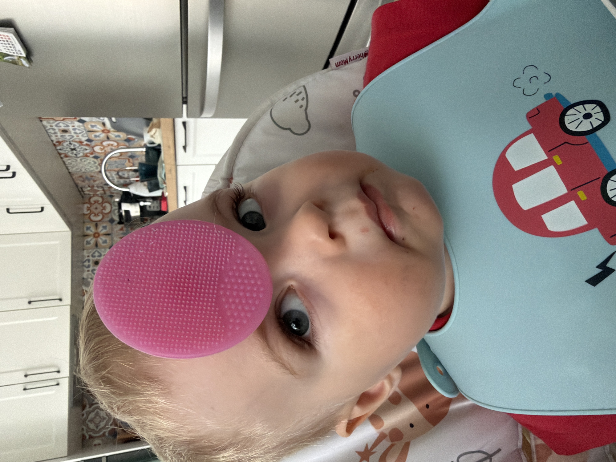 A baby wearing a light blue bib with a red car design is lying down. A pink, circular object covers a portion of the baby's forehead. The location is not discernible from the image, and therefore I cannot ascertain if it is in the city of Votkinsk, Russia. The background shows kitchen cabinets, a tiled backsplash, and a metallic object. The baby is the primary subject of the image.