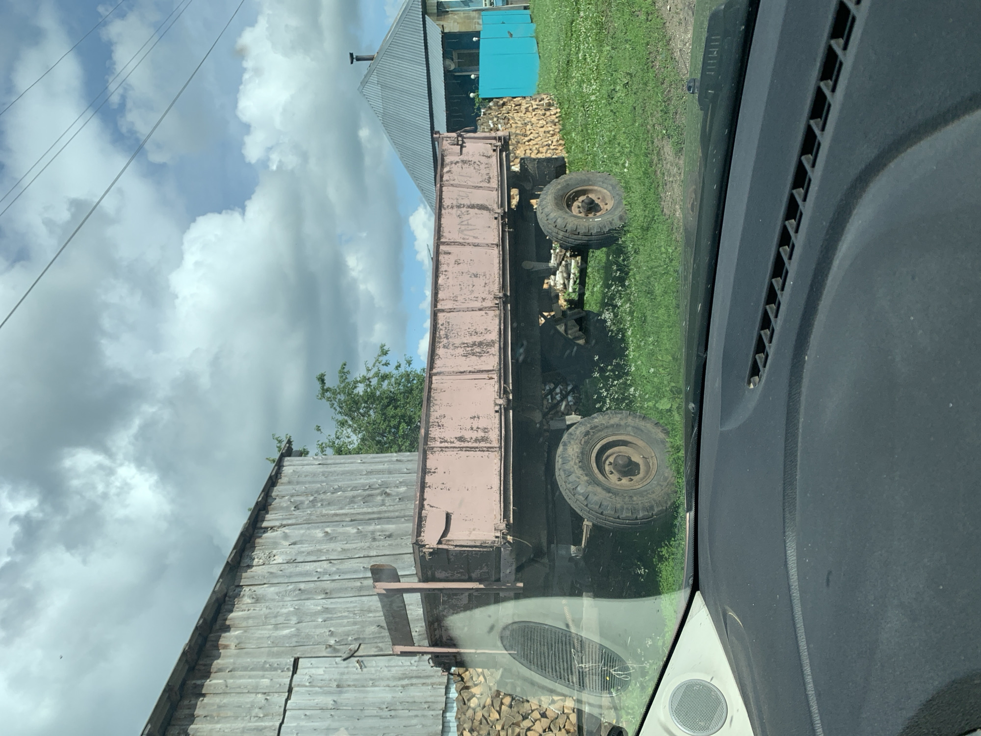 This is a medium-angle, outdoor shot of a dilapidated trailer parked in front of a wooden structure on a cloudy day. The trailer, with peeling pink paint, has two large, worn tires and a metal bed. To the left of the trailer, a stack of firewood is visible against the wooden building. Beyond the trailer, a blue structure and a grey-roofed building are present. The ground is covered in grass and some wildflowers, with a dirt track nearby. The image appears to be taken from inside a vehicle, with a portion of the dashboard and a speaker visible in the foreground. The scene is set in the rural environment of Barsai, Russia.
