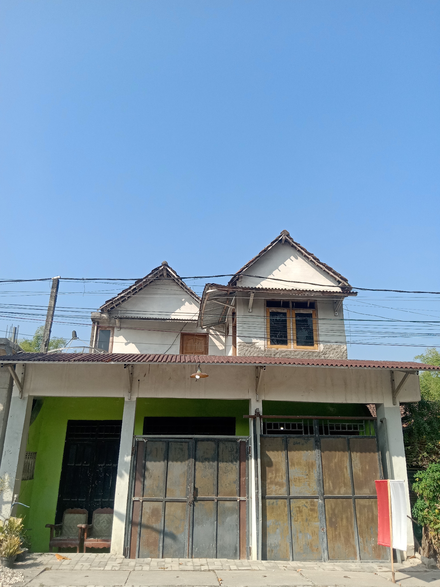 The image shows the exterior of a two-story house with a light-colored facade and a corrugated metal roof. The house has two distinct rooflines, each with a triangular gable. The upper story features two windows with wooden frames, one with glass panes and the other with what appears to be wooden shutters. Below the upper story, there is a covered porch area with a light green painted wall and two large, weathered metal gates that span the width of the property. To the left of the gates, two old armchairs are visible on the paved ground in front of the green wall. Several power lines are strung across the upper portion of the image, with a concrete utility pole on the left. To the right of the house, there is dense green foliage. A small flag with red and white stripes is mounted on a pole to the right of the rightmost gate. The sky is a clear, bright blue. The scene is likely in Tegalkodo, Indonesia, given the architectural style and vegetation.