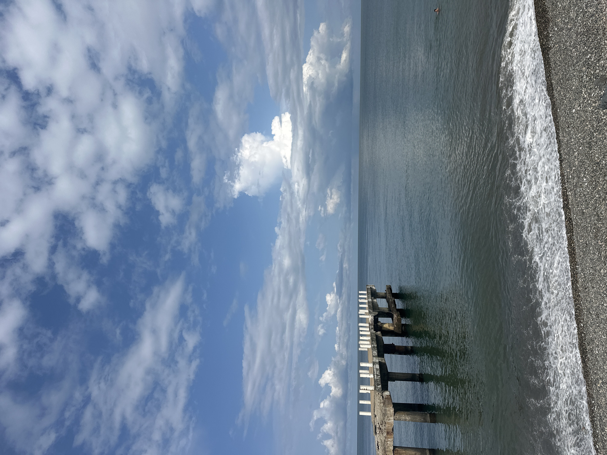 An abandoned concrete pier extends from a pebbled shore into a calm, dark blue sea under a sky with scattered white clouds. The pier, partially in disrepair with exposed rebar, is supported by multiple concrete pillars and features white posts along its edge. The water near the shore has gentle waves lapping against the pebbles. In the distance, a single person is swimming in the sea. The scene is likely located in Batumi, Georgia, a coastal city known for its piers and Black Sea coastline.