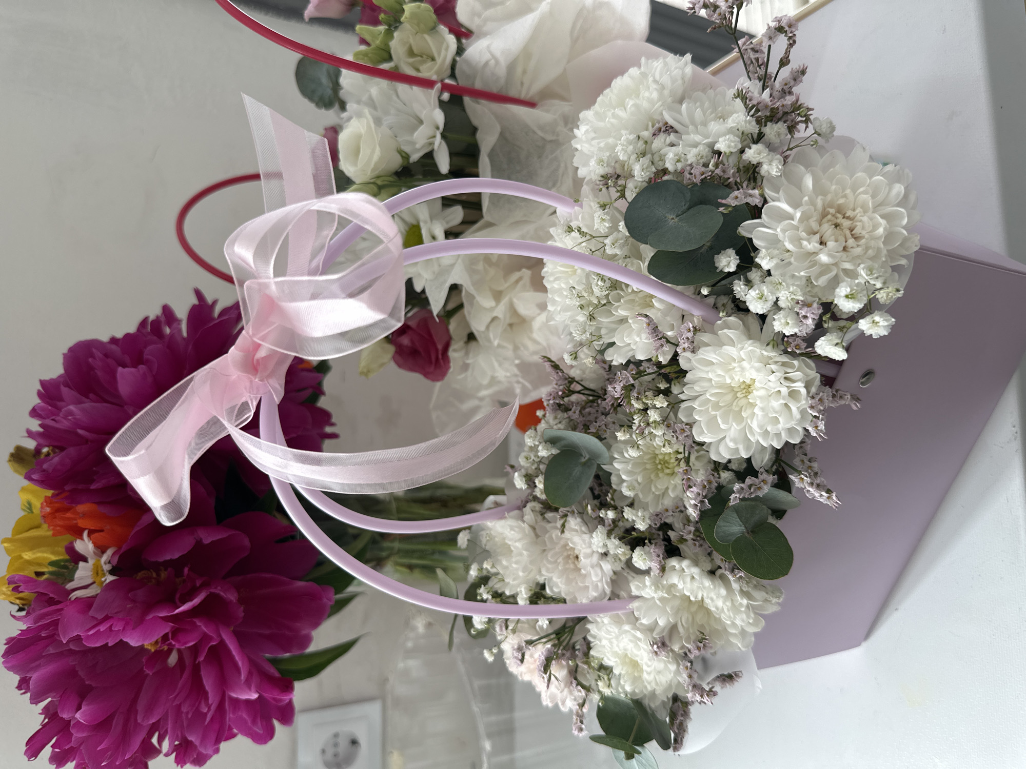 In an indoor setting with a neutral, light-colored background, a light pink floral box is filled with a dense arrangement of white chrysanthemums and smaller white flowers, accented with sprigs of delicate light purple flowers and eucalyptus leaves. A sheer pink ribbon is tied in a bow over two light pink wire handles that arch out of the box. In the background, a vase holds a bouquet of vibrant magenta peonies and yellow flowers. The image appears to be taken from a slightly elevated angle, with the floral box positioned in the foreground on a white surface.