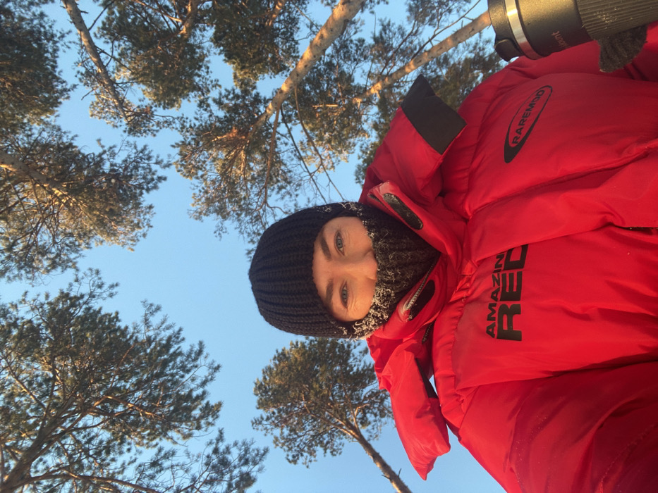 A low-angle outdoor selfie captures an individual, likely female, in Ust-Ilimsk, Russia, under a clear blue sky. The subject wears a black ribbed knit beanie and a dark grey knit neck gaiter covering the lower face, with visible frost crystals on the fibers. Her eyes are blue, and her expression is neutral. She is dressed in a bright red puffer jacket; black text "AMAZING RED" is visible on the left chest, and "RAREMOD" with an oval logo is on the right chest. She holds a dark grey travel mug or thermos, partially obscuring the brand "vount," in her right hand. The background consists of numerous tall pine trees with green needles and brown branches, silhouetted against the bright sky, suggesting a natural, possibly forested, outdoor environment in cold weather conditions.