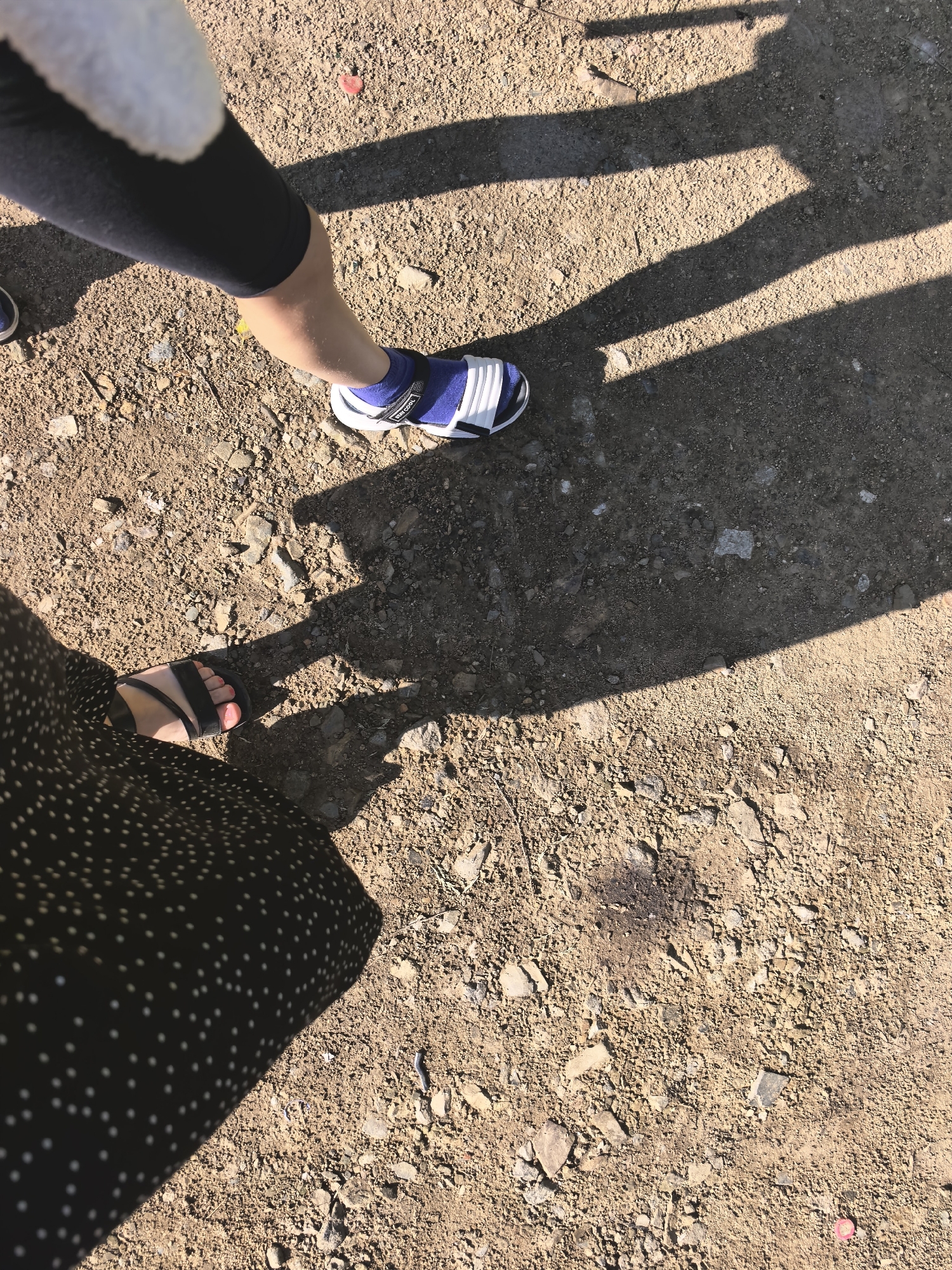 The image is an overhead shot of two people's legs and feet standing on a gravel path. The person on the left is wearing a black polka-dot skirt and black sandals. The person on the right is wearing a white and blue sandal with blue socks and black leggings. Shadows from an unseen object fall across the scene, creating a striped pattern on the ground. The ground is covered in small stones and dirt. A small red object is visible on the ground in the upper right portion of the image.