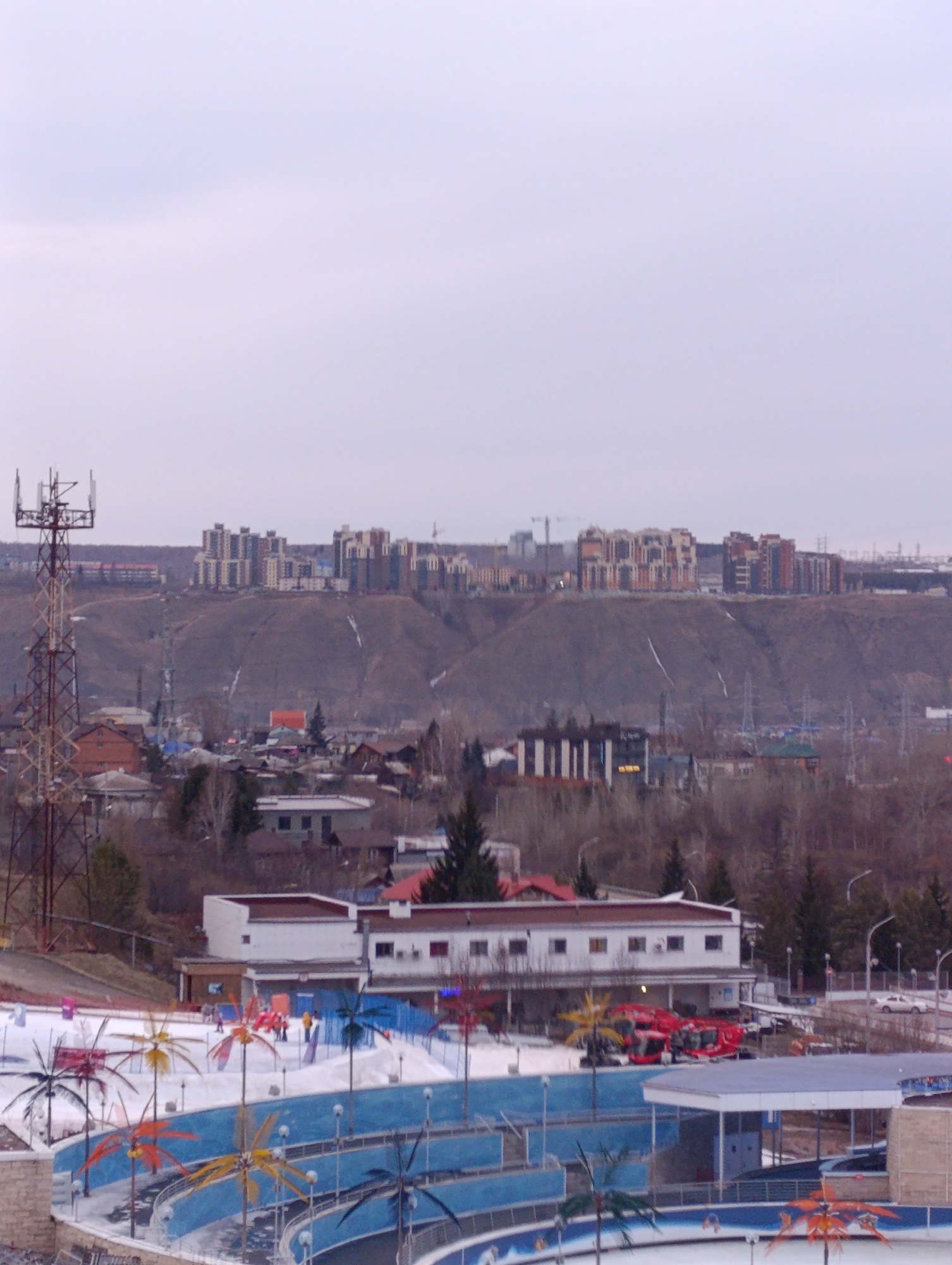 The image captures a cityscape in Krasnoyarsk, Russia, featuring a foreground with an artificial aquatic park. The pools are painted blue, and there are decorative artificial palm trees scattered throughout the area.
In the middle ground, there is a white, low-rise building with a flat roof. Behind the building, a dense array of trees and a residential area are visible, indicating a mix of urban and natural environments.
In the background, a tall hill dominates the skyline, and on the hilltop, there are a number of high-rise residential buildings. To the left of the hill is a communications tower.
The weather appears overcast, with a pale sky providing a soft light over the scene.